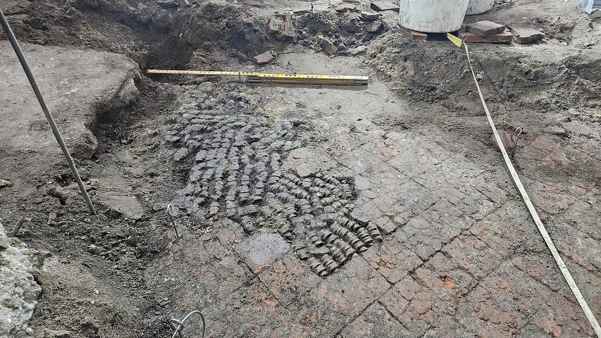 Archeologische opgraving met stenen vloer en mozaïekpatroon, omgeven door zand en aarde. Meetlinten zichtbaar op de achtergrond.