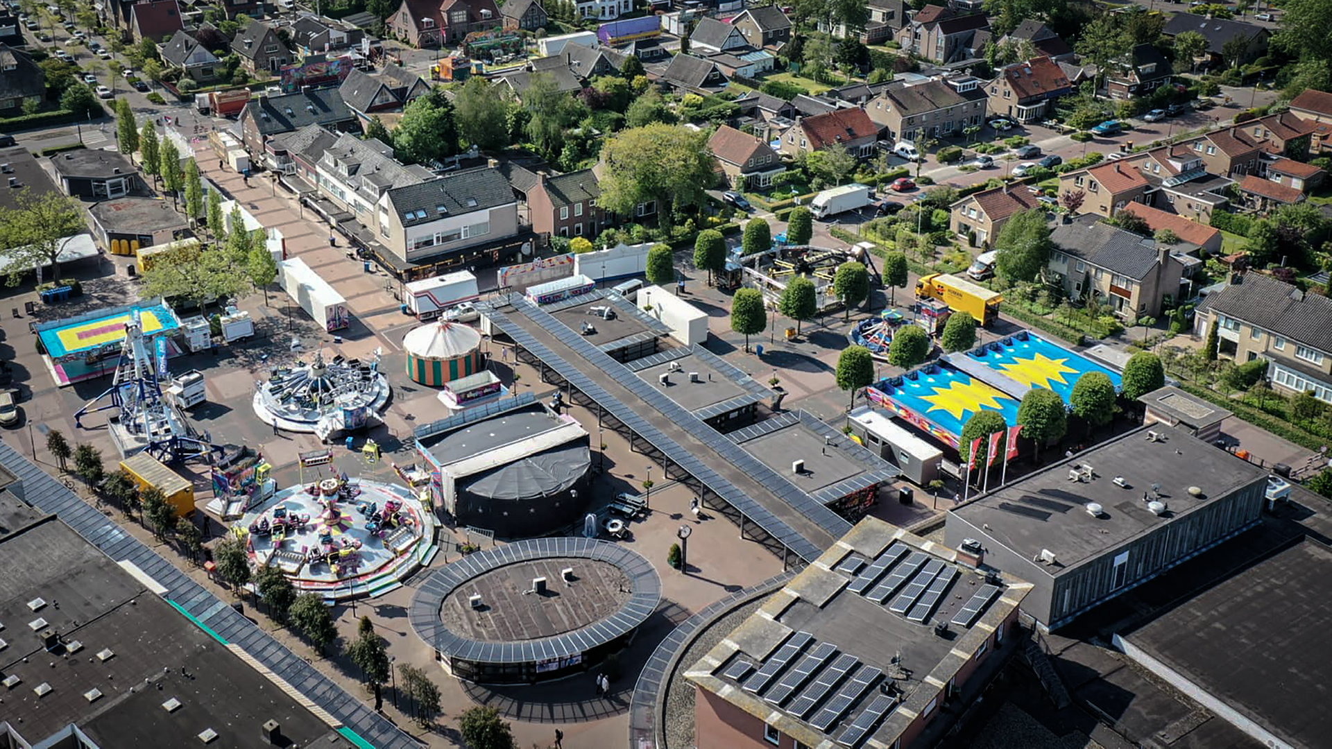 Luchtfoto van een kermis in een woonwijk met verschillende attracties, kraampjes en omliggende huizen.