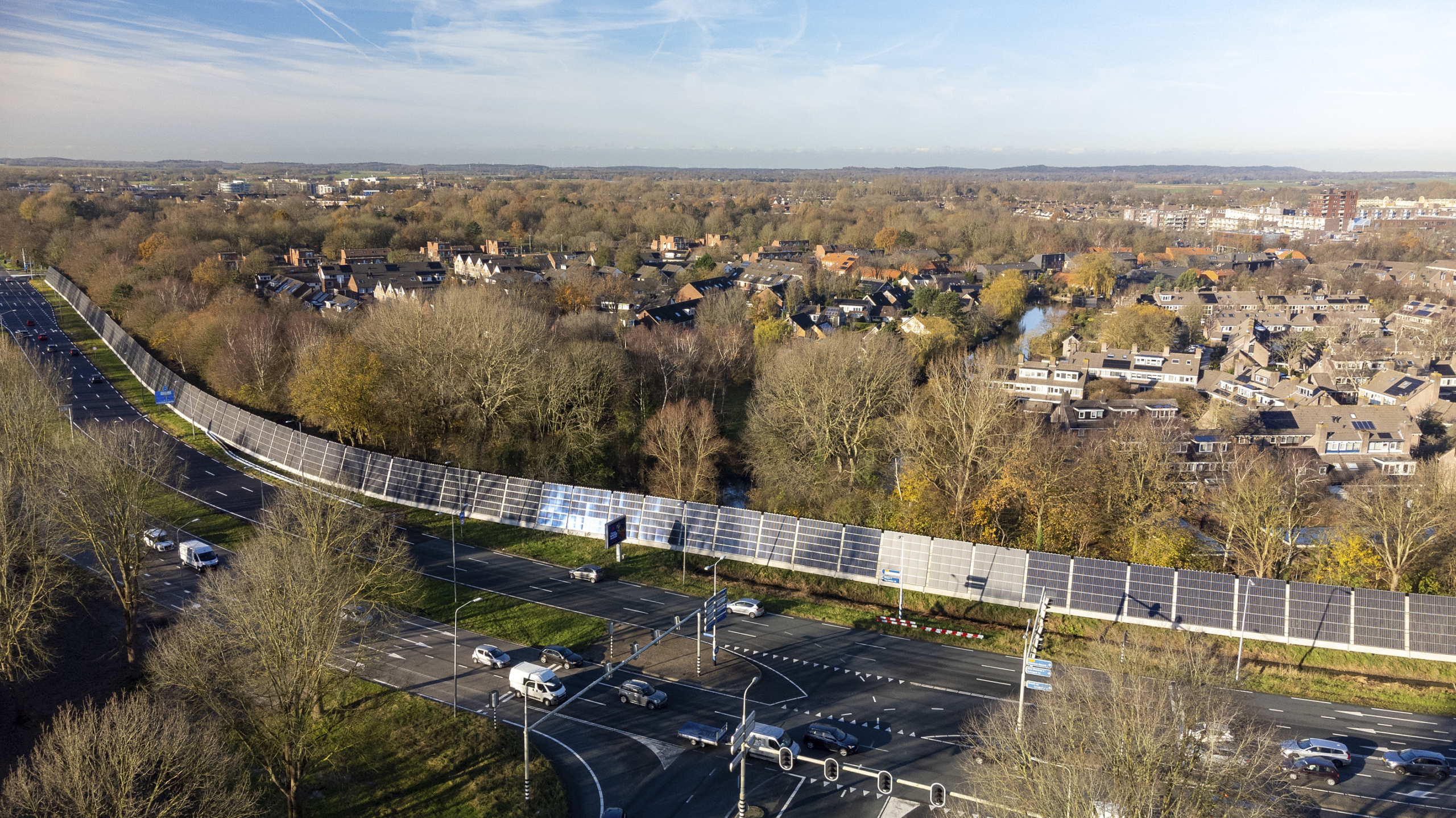 Luchtfoto van een weg naast een woonwijk, met een rij zonnepanelen tussen de weg en de huizen, omgeven door bomen in herfstkleuren.