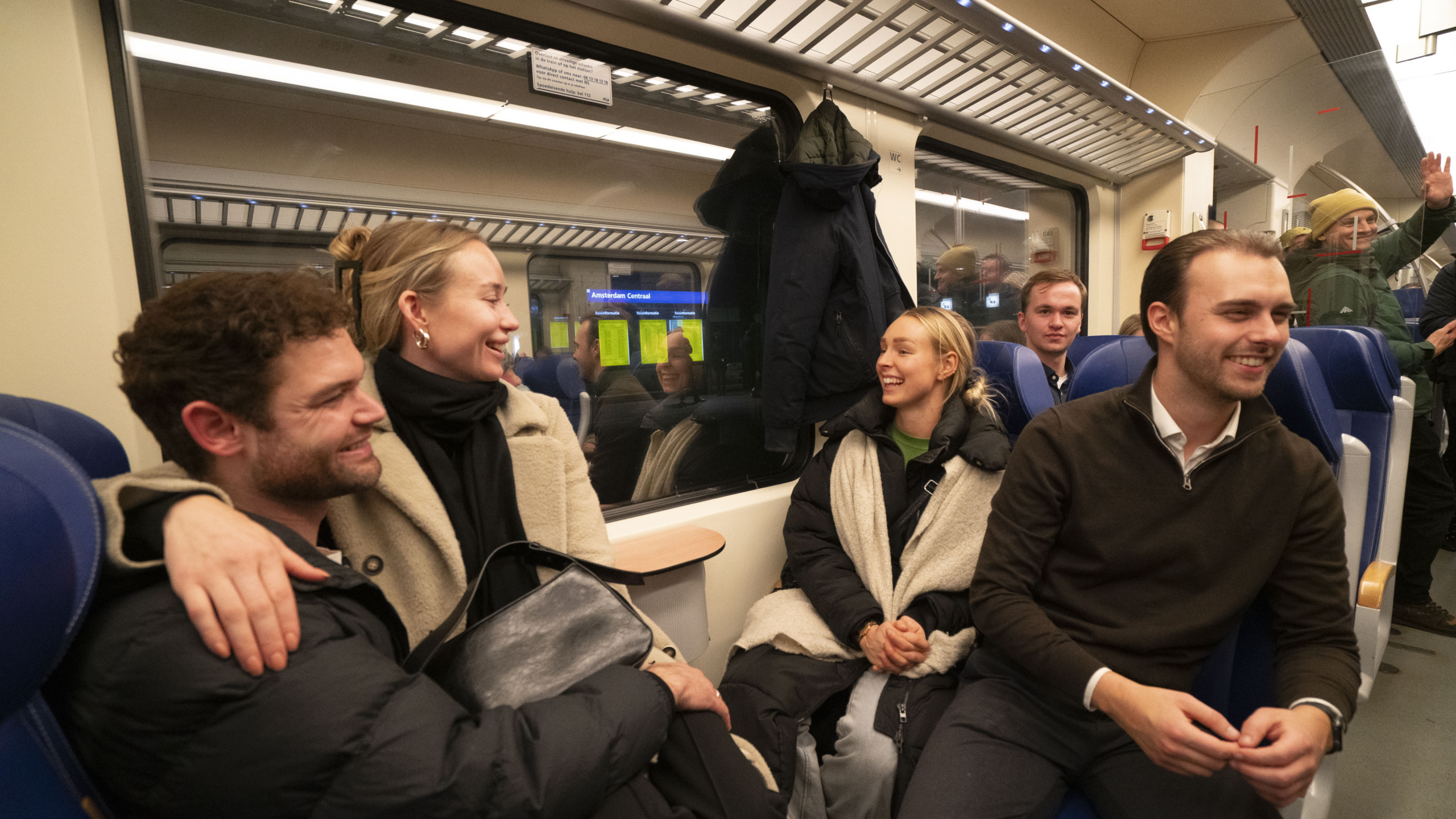 Groep mensen glimlachend en pratend in een trein, zittend op blauwe stoelen. Er hangt een jas aan de kapstok en op de achtergrond zie je een station.