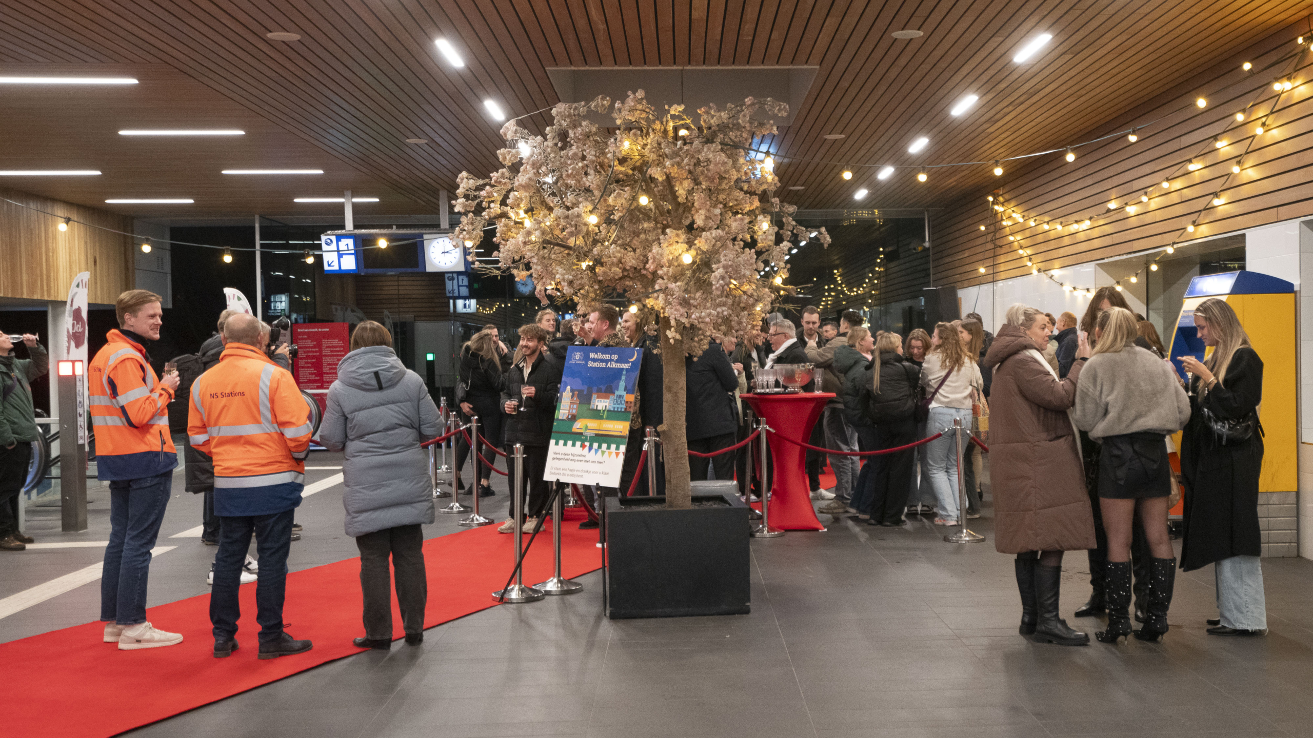 Mensen verzamelen zich bij een versierd station met rode loper en feestelijke verlichting.