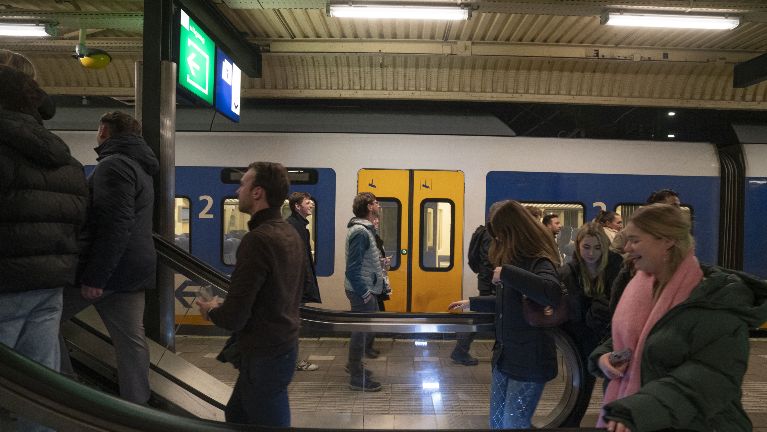 Mensen op een treinstation, sommigen op de roltrap en anderen lopen langs een stilstaande trein.