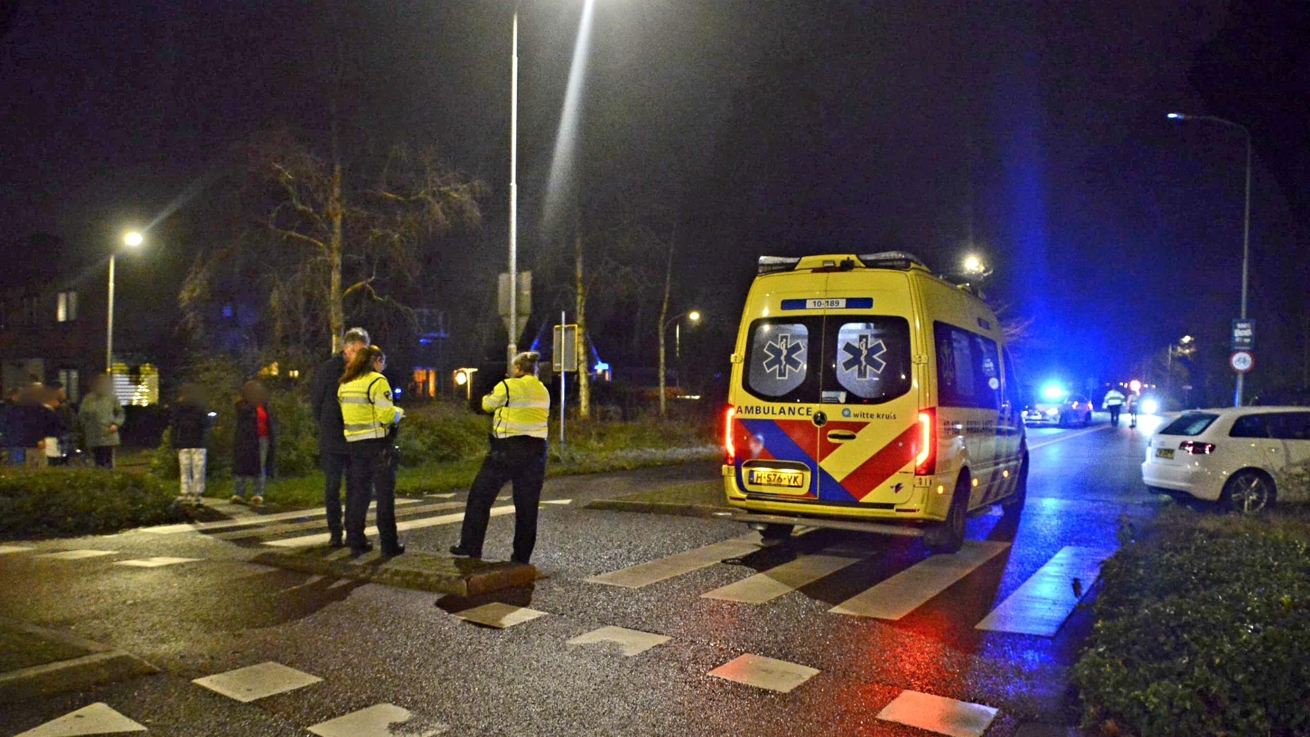 Ambulance op straat bij nacht, met twee politieagenten en enkele omstanders.