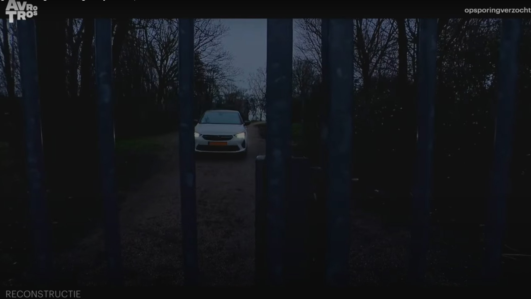 Een witte auto staat geparkeerd achter een hek aan het einde van een onverharde weg, omgeven door bomen in de schemering.