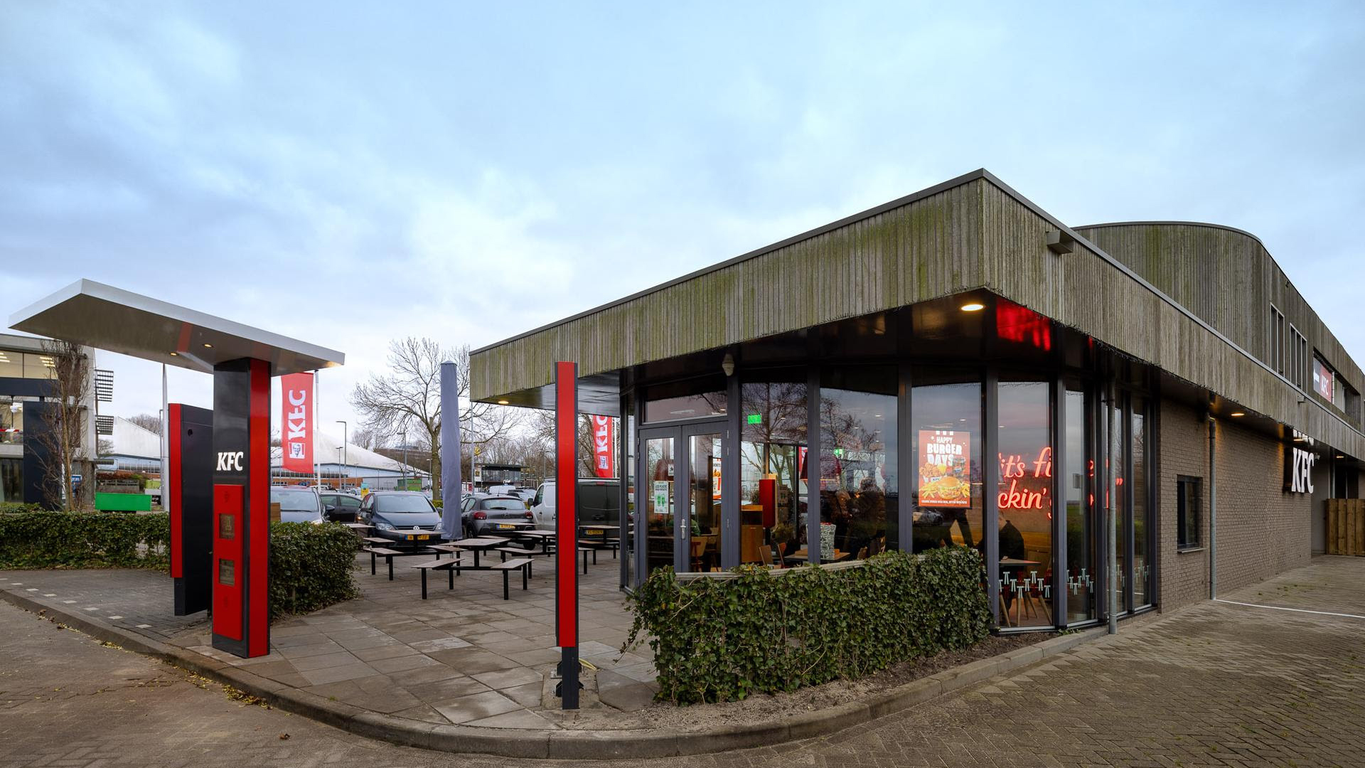 Buitenzijde van een KFC-restaurant met parkeerplaatsen en een drive-thru-zuil op een bewolkte dag.