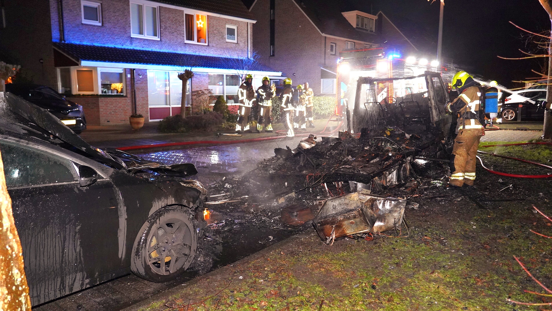 Brandweermannen blussen een uitgebrand voertuig bij nacht voor een huis met kerstverlichting.