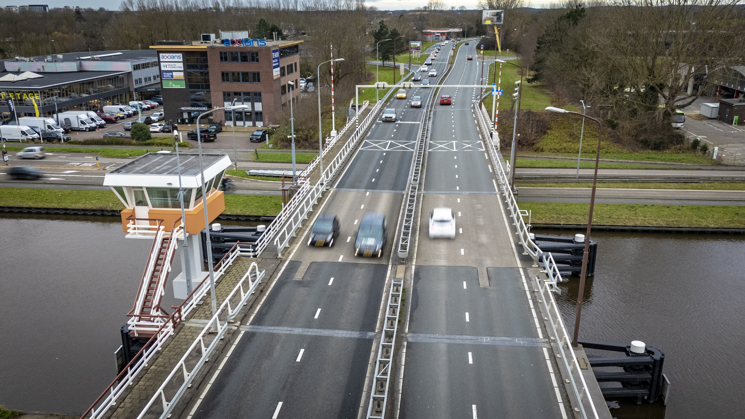 Autos rijden over een brug boven een kanaal, met een kantoorgebouw en parkeerplaats aan de linkerkant.