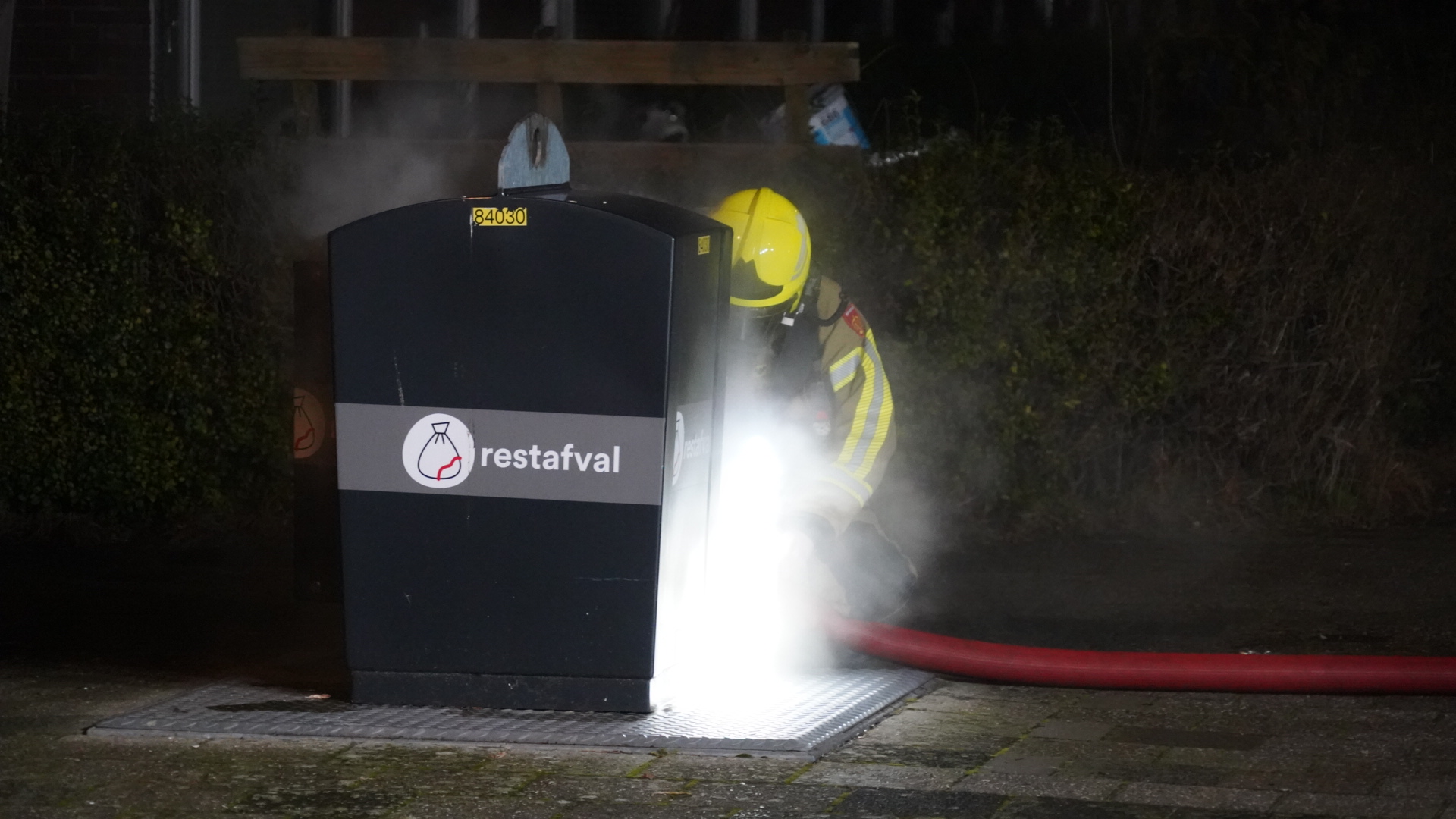 Brandweerman blust brand in container voor restafval met brandslang.