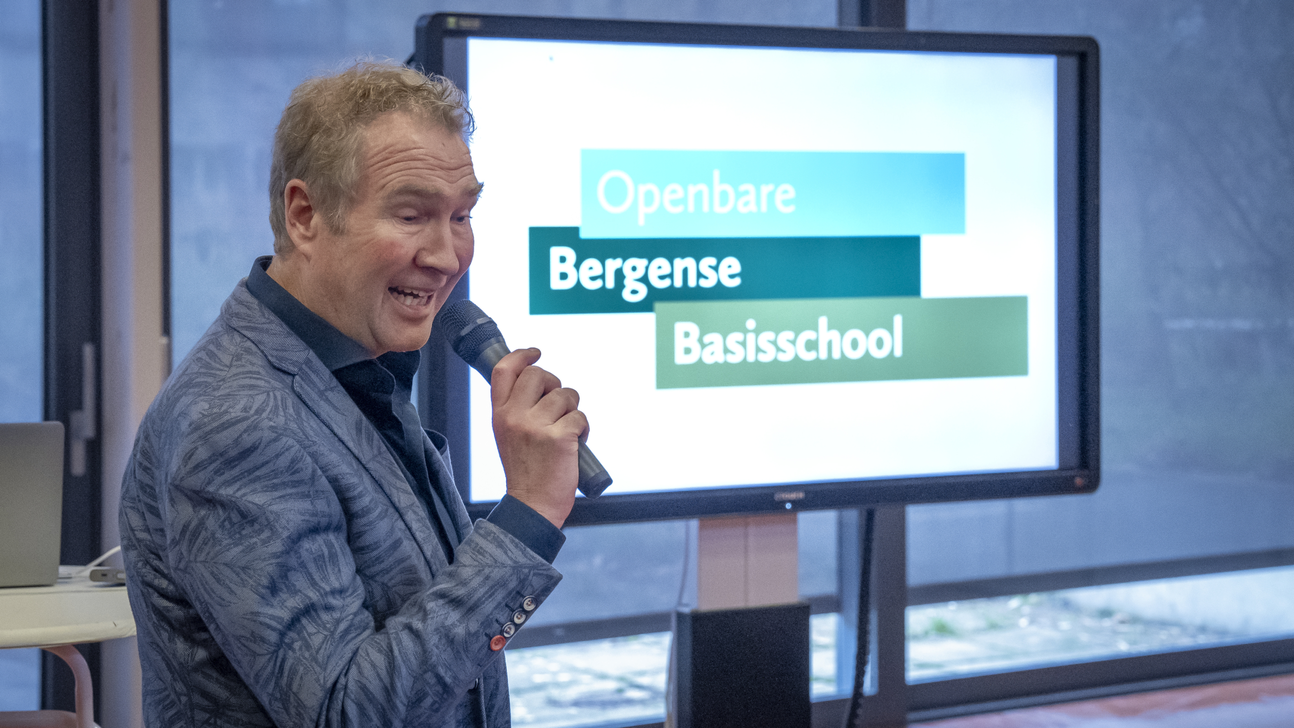 Man met microfoon presenteert voor scherm met tekst "Openbare Bergense Basisschool".