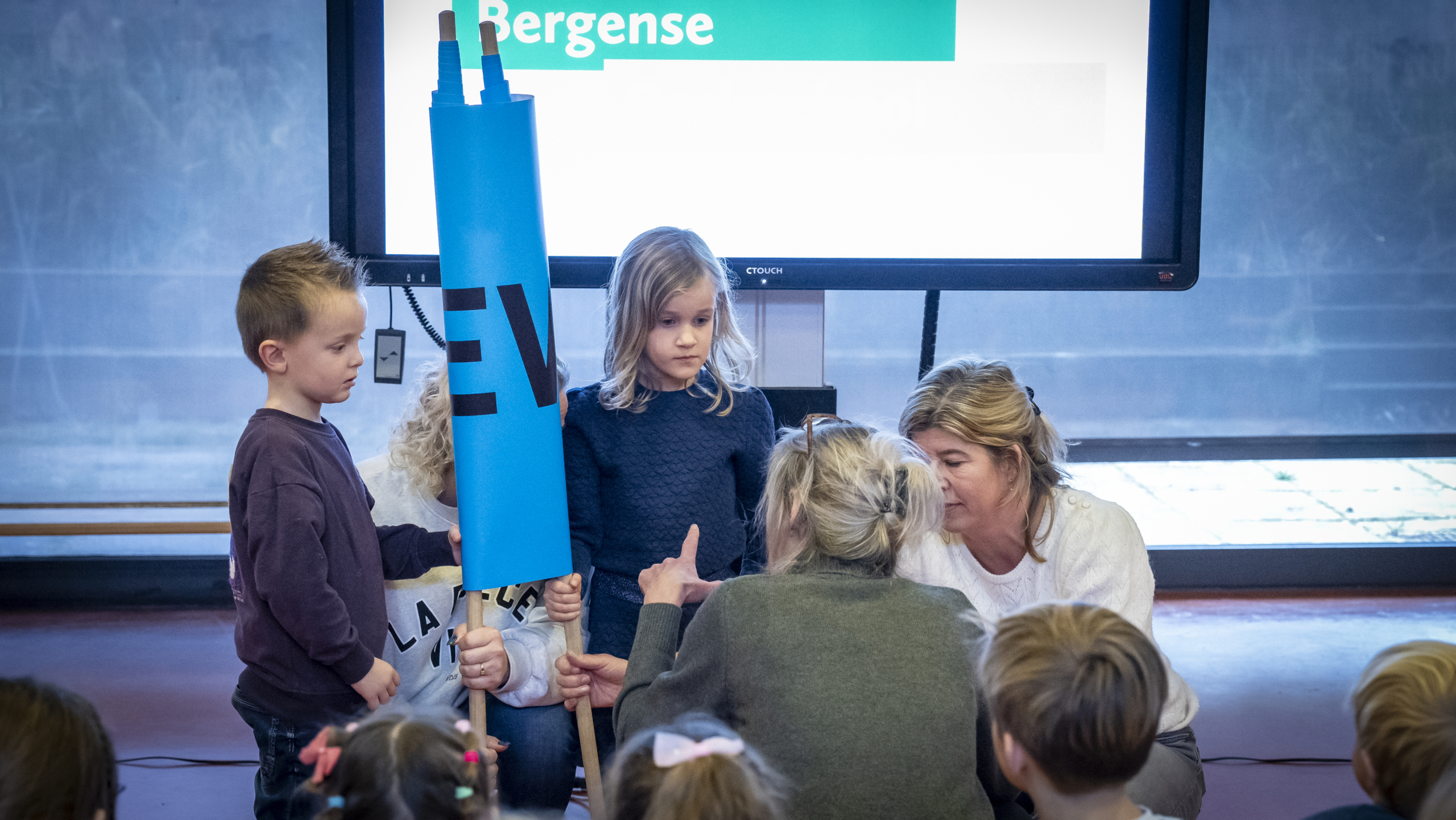 Kinderen en volwassenen zijn bezig met een activiteit voor een scherm; een kind houdt een blauw bord vast.