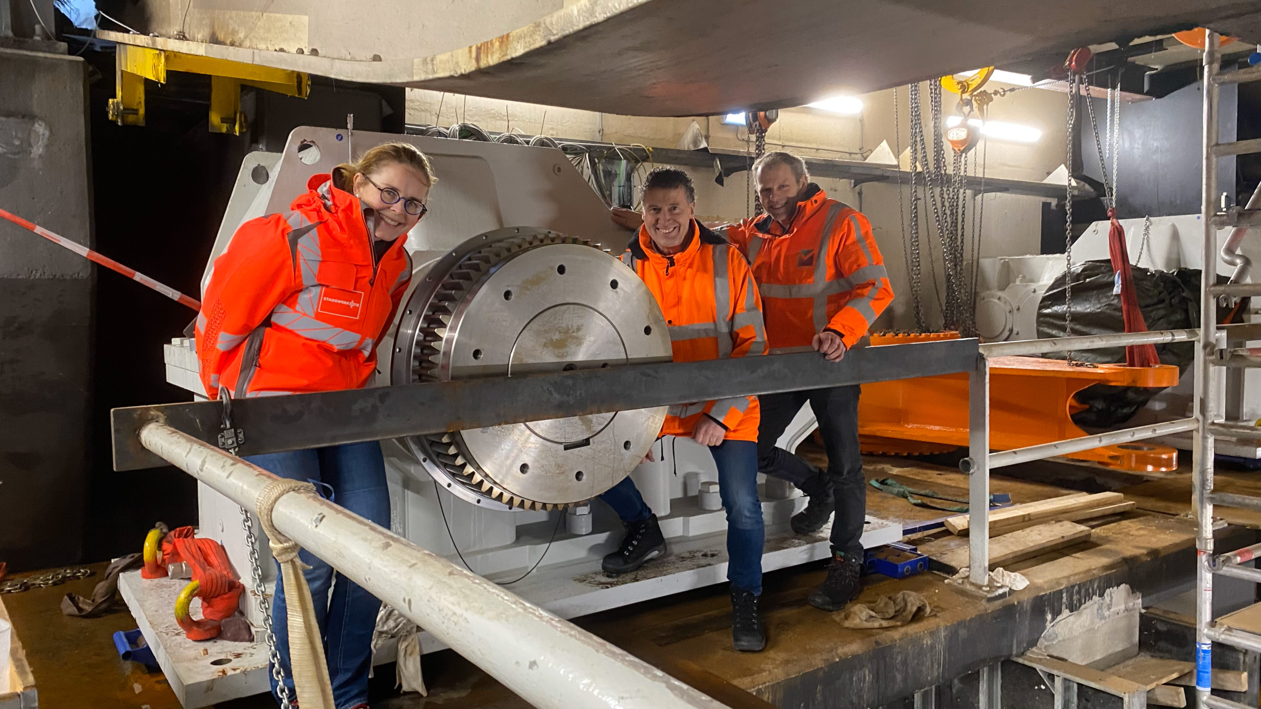 Drie personen in oranje veiligheidsjassen poseren naast een grote industriële tandwielmachine op een bouwlocatie.