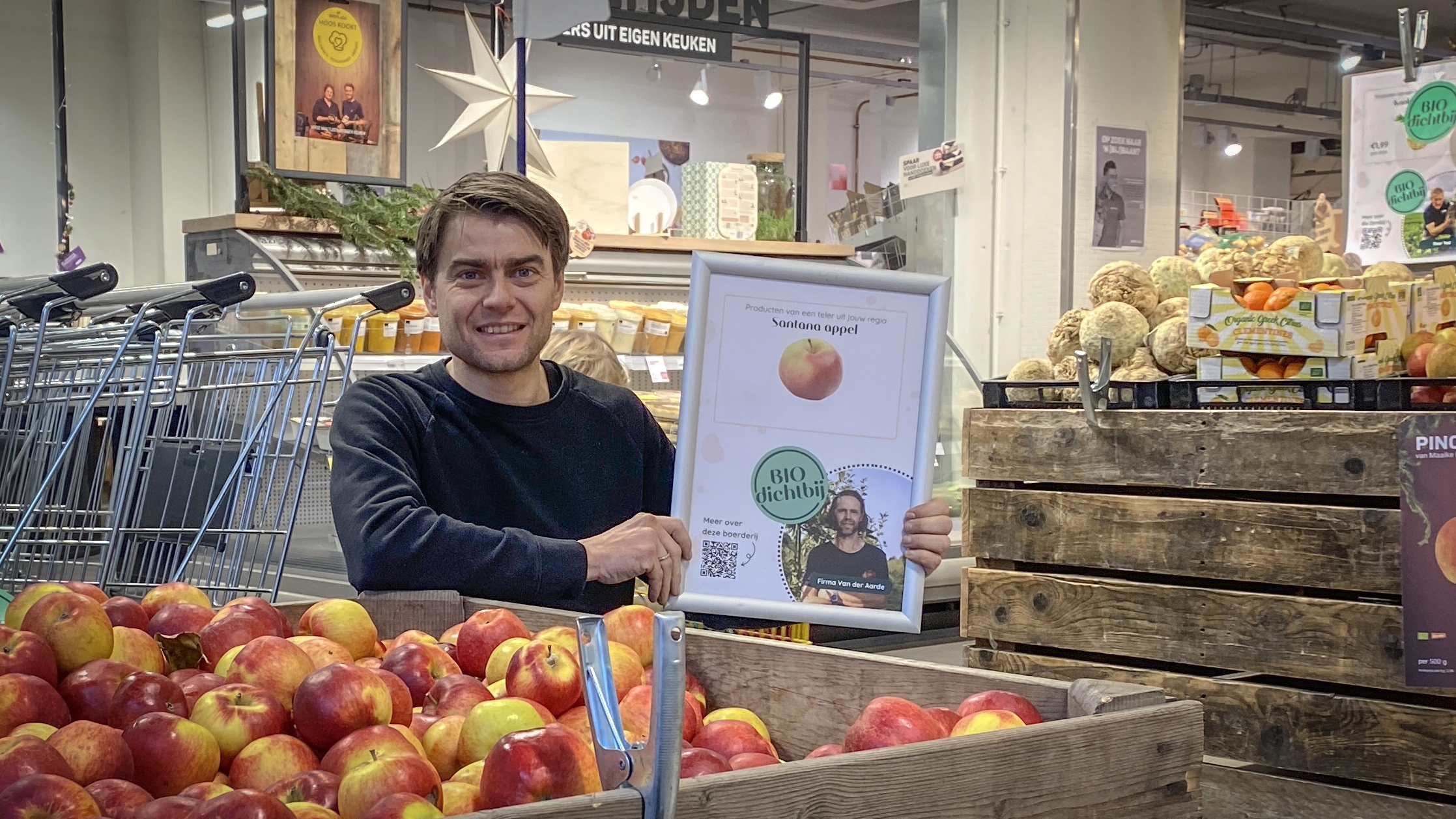 Man in supermarkt houdt een bord vast bij een display met Santana appels.