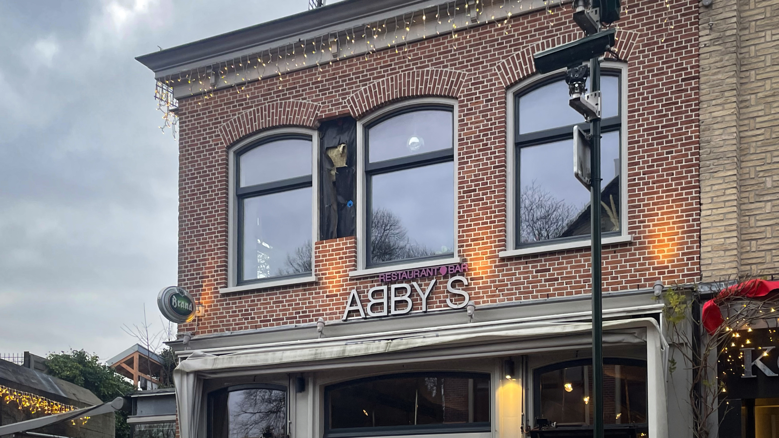 Bakstenen gebouw met twee ramen, kleine lichtjes aan de rand van het dak en een bord met de tekst "Restaurant | Bar ABBY'S" boven de ingang.