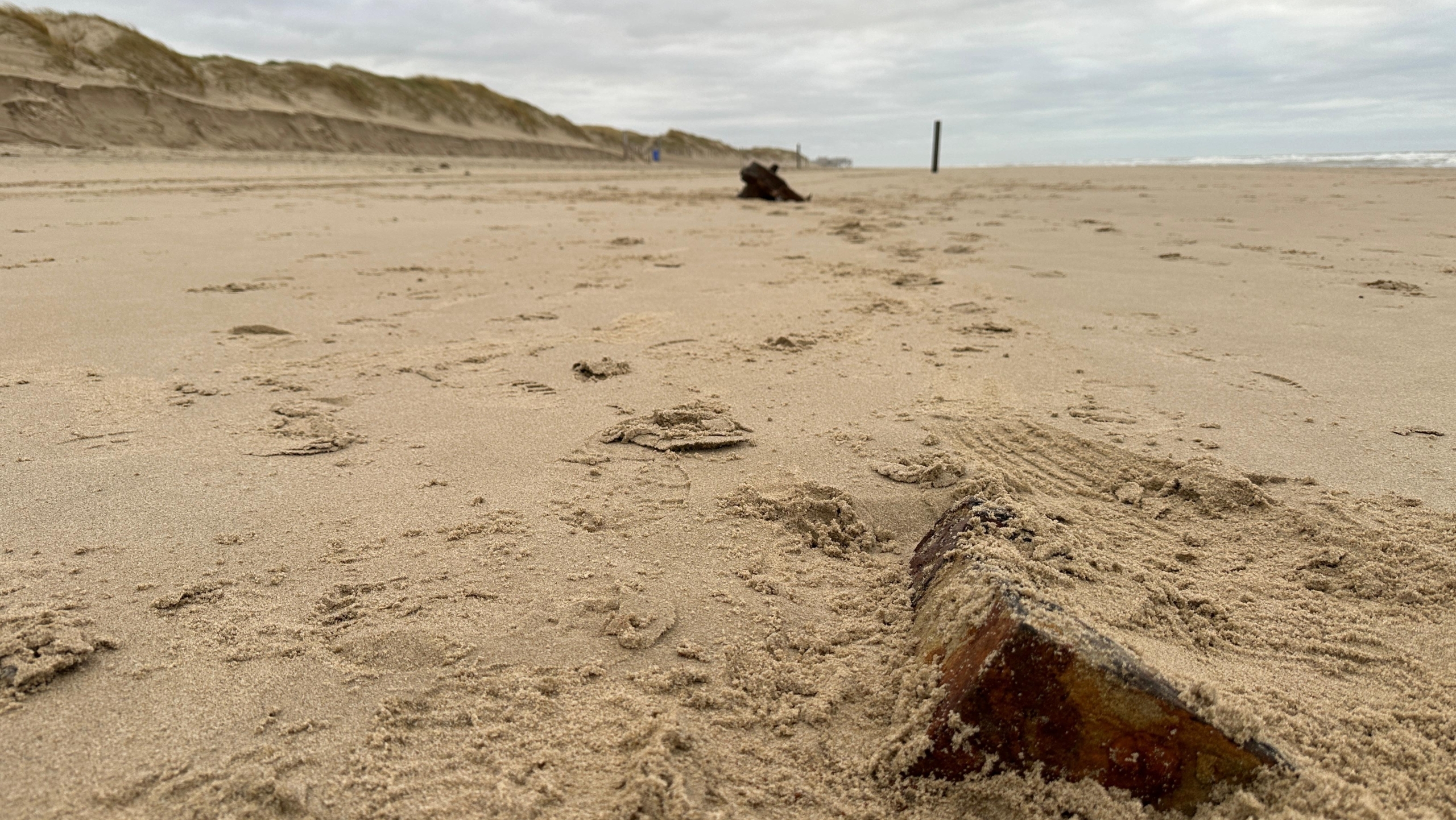 Zandstrand met een scherf baksteen op de voorgrond en duinen op de achtergrond onder een bewolkte hemel.