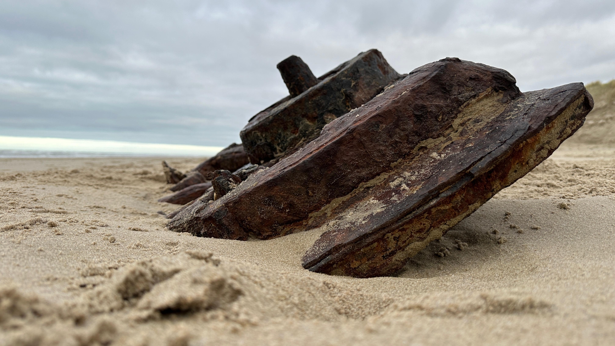 Roestig metaal gedeeltelijk begraven in het zand op een strand onder een bewolkte lucht.