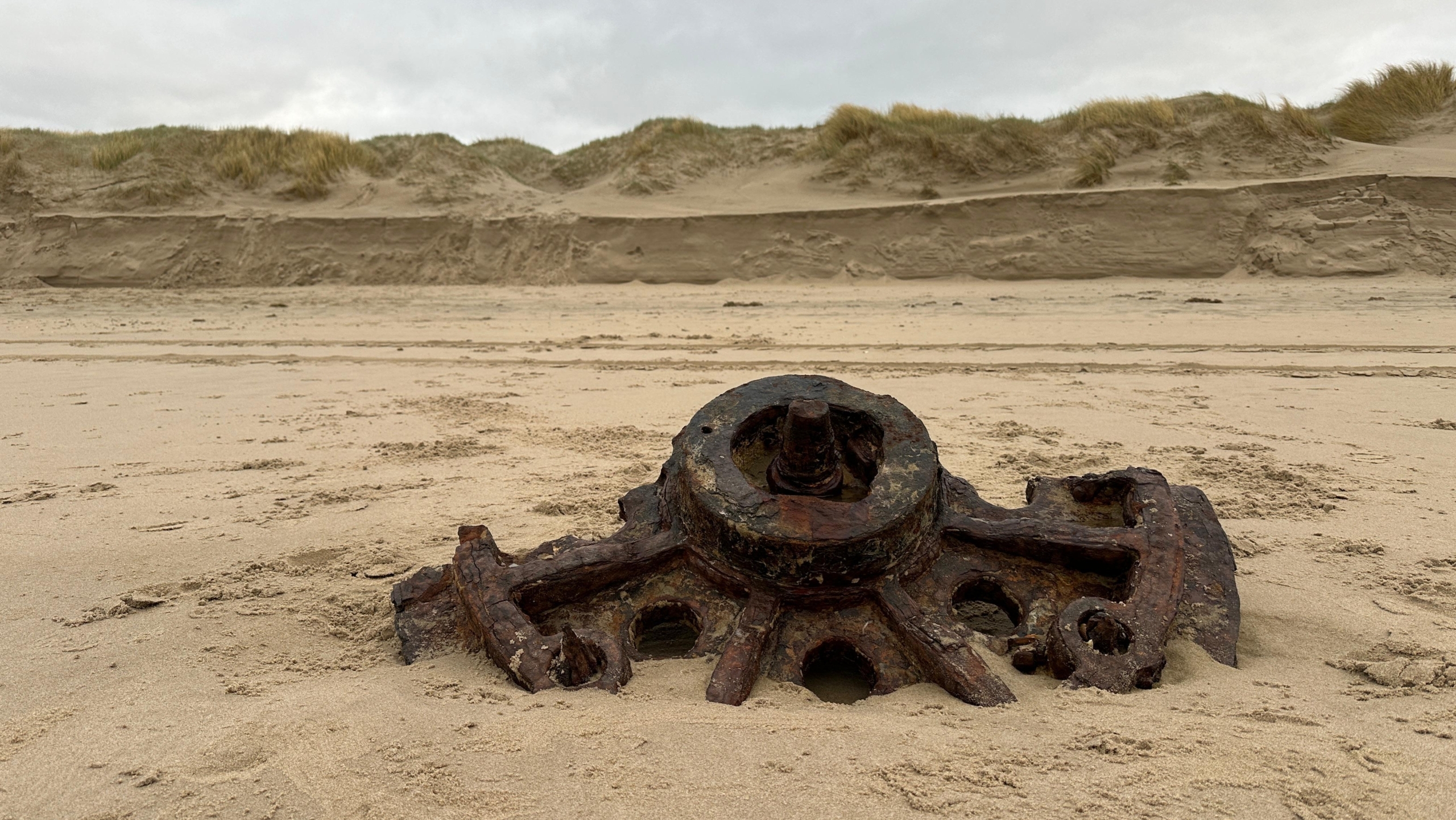 Roestig mechanisch onderdeel half begraven in zand op het strand, met duinen op de achtergrond.