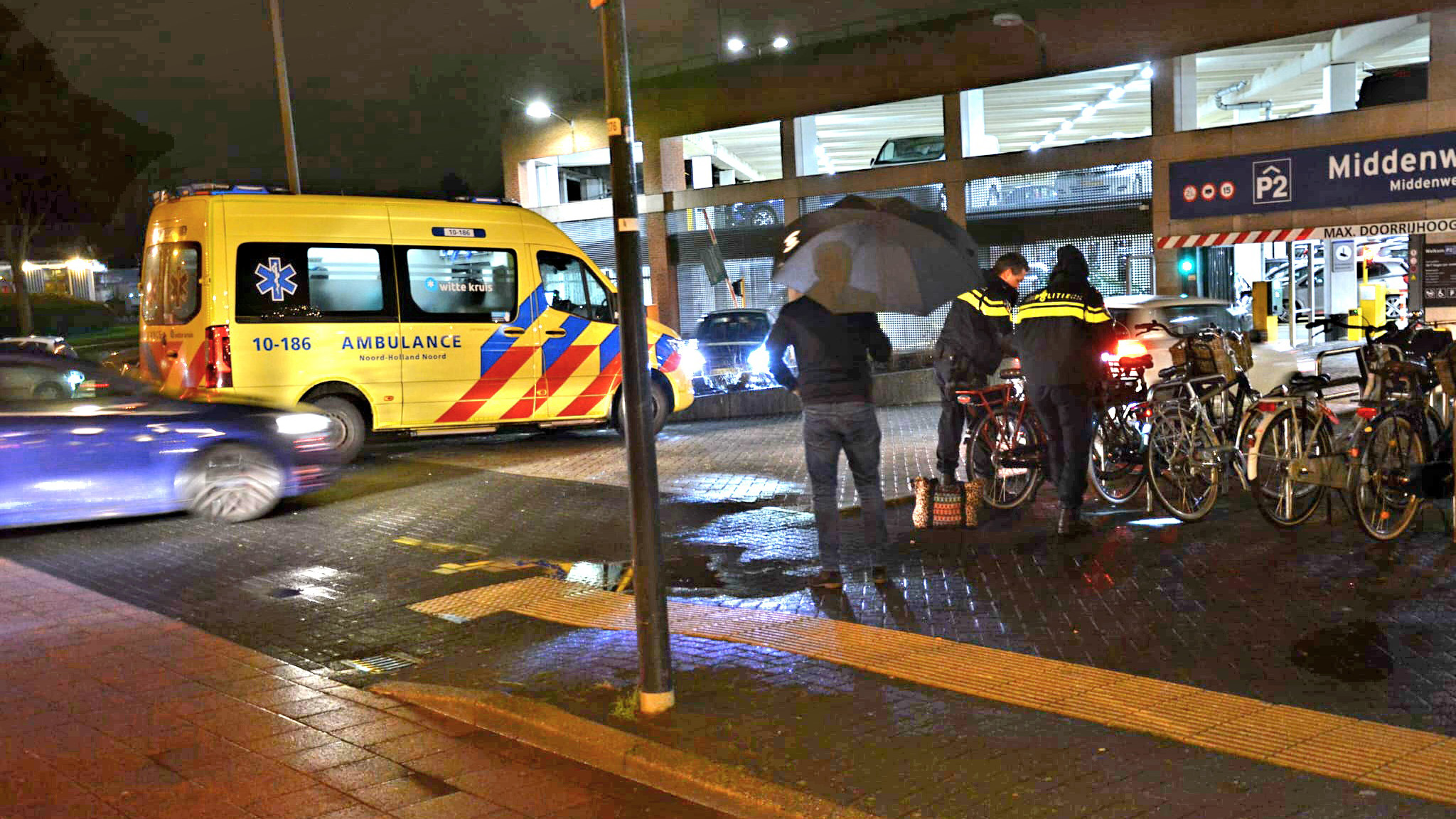 Een gele ambulance en een politieagent bij een fietsenstalling op een regenachtige avond.