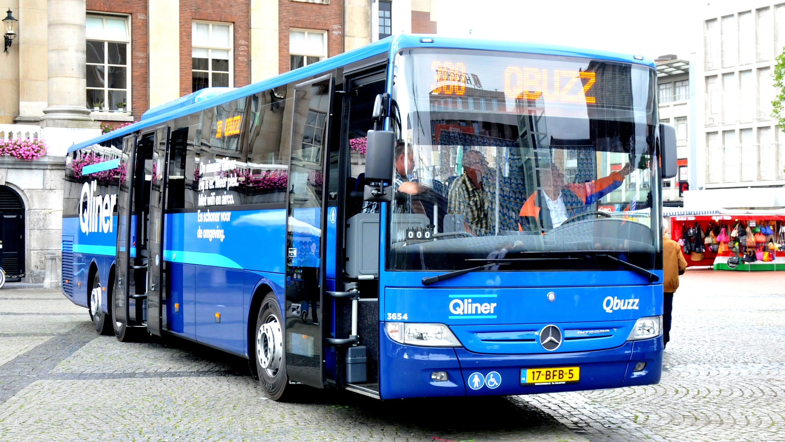 Blauwe Qliner-bus van Qbuzz op een plein met marktkramen op de achtergrond.