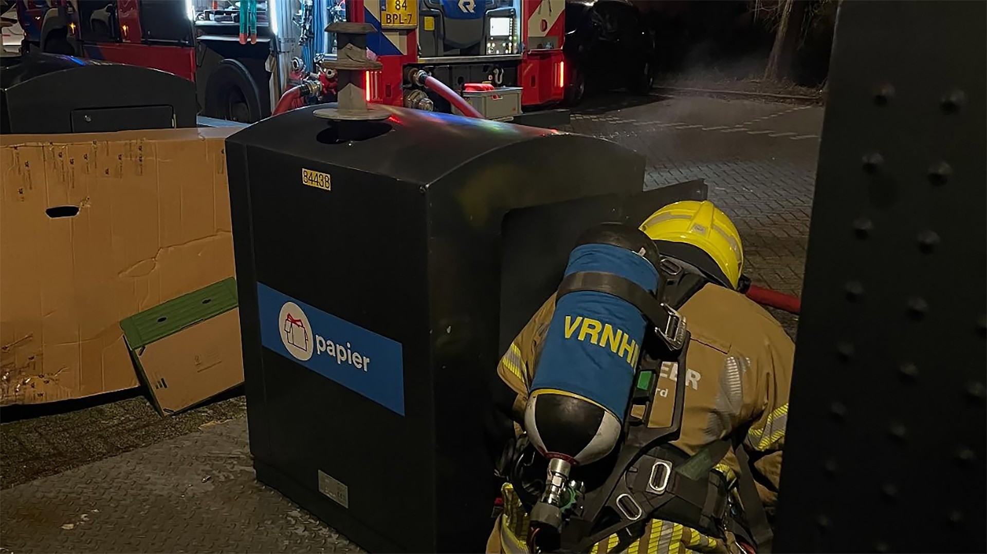 Brandweerman in beschermende uitrusting onderzoekt een papiercontainer naast een brandweerwagen.