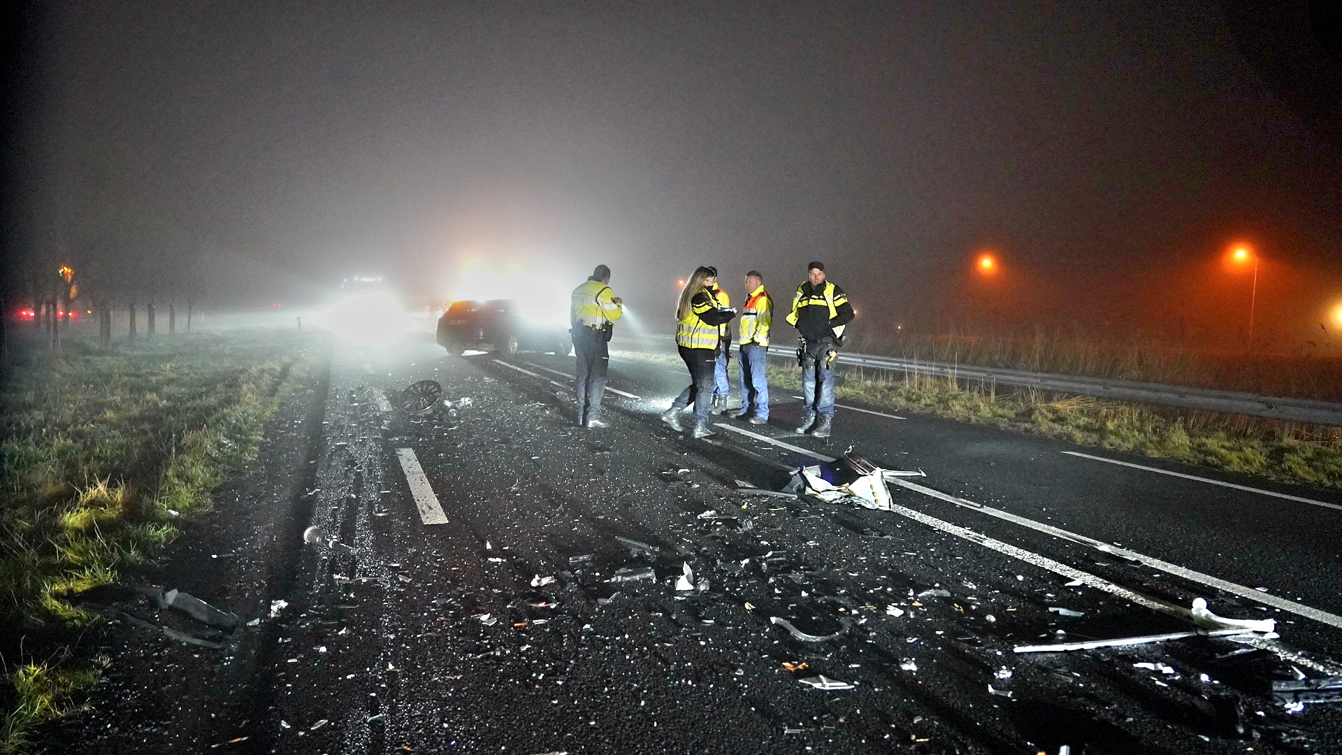 Politie en hulpverleners op een mistige weg na een verkeersongeluk met wrakstukken op het asfalt.