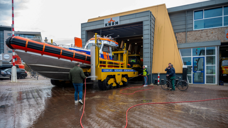 Een reddingsboot op een gele trailer wordt buiten een gebouw van de KNRM gewassen. Meerdere mensen staan om de boot heen, waaronder een persoon met een fiets.