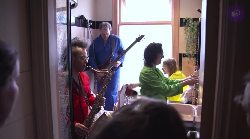 Mensen spelen muziek in een kleine badkamer, waaronder een saxofonist en gitarist.