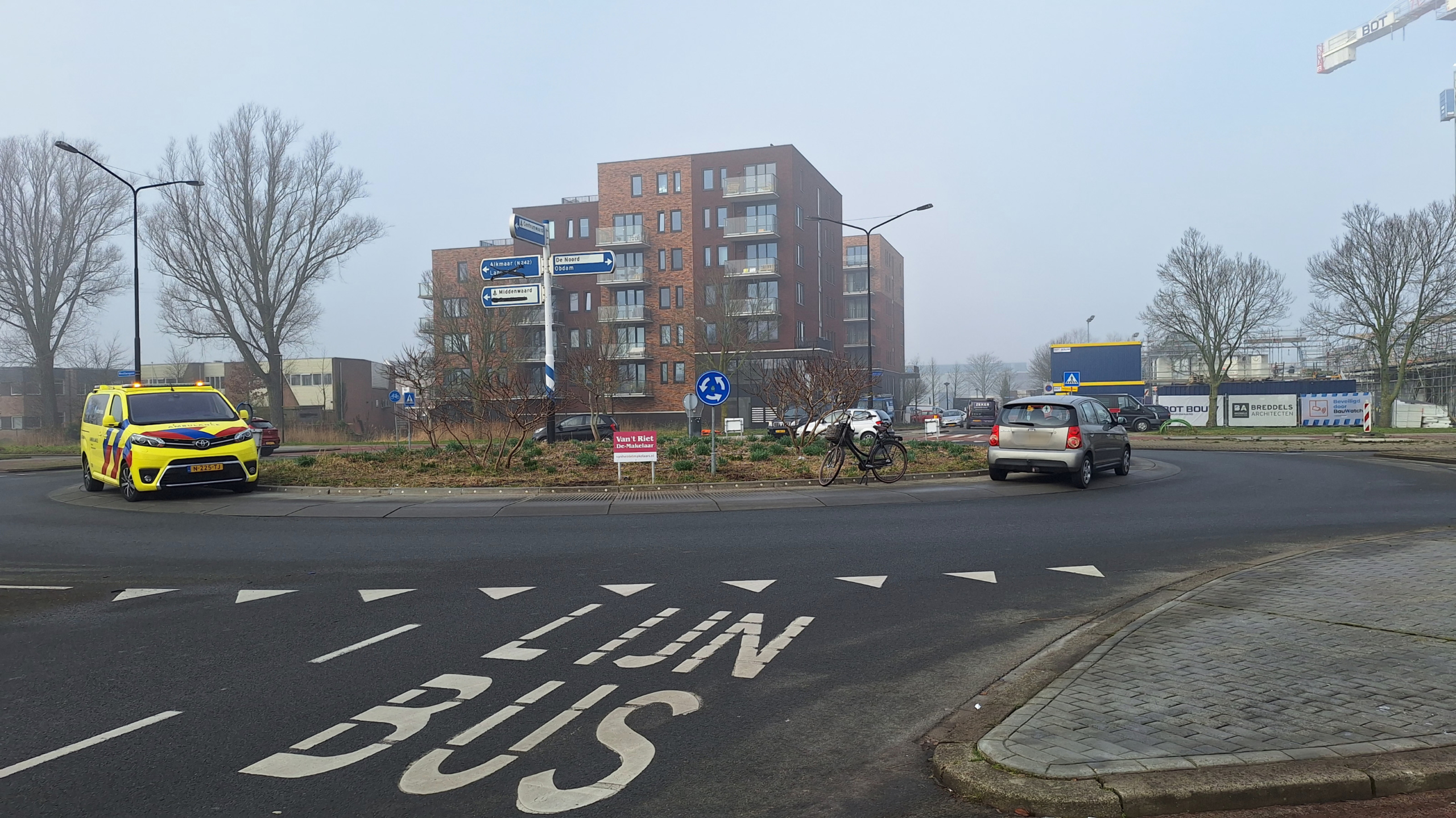 Een rotonde met een geel hulpdienstvoertuig en een grijze auto, omgeven door gebouwen, verkeersborden en een fiets tegen een paal.