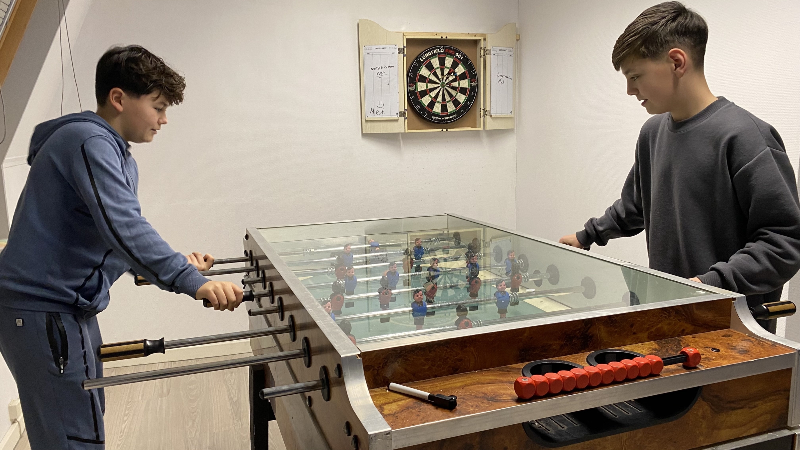 Twee jongens spelen tafelvoetbal in een kamer met een dartbord aan de muur.