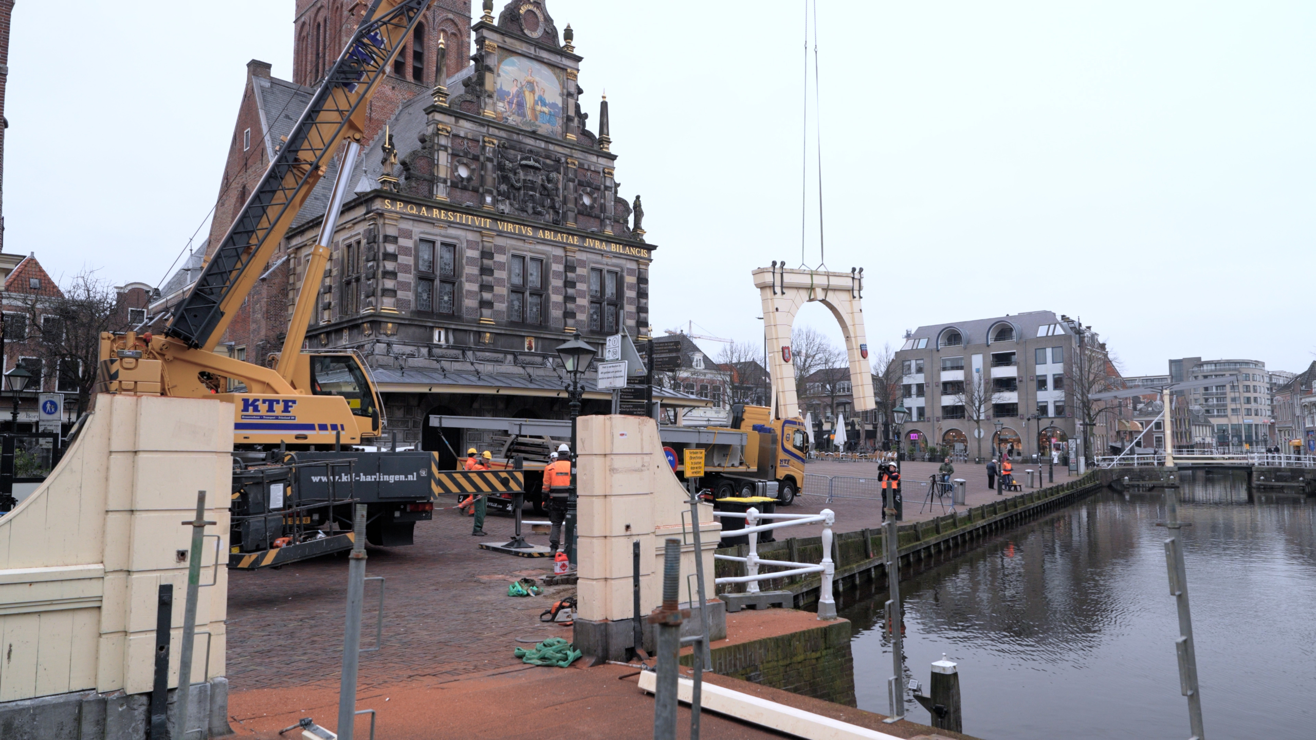 Bouwplaats met kraan en arbeiders bij een historische brug en gebouw aan het water.