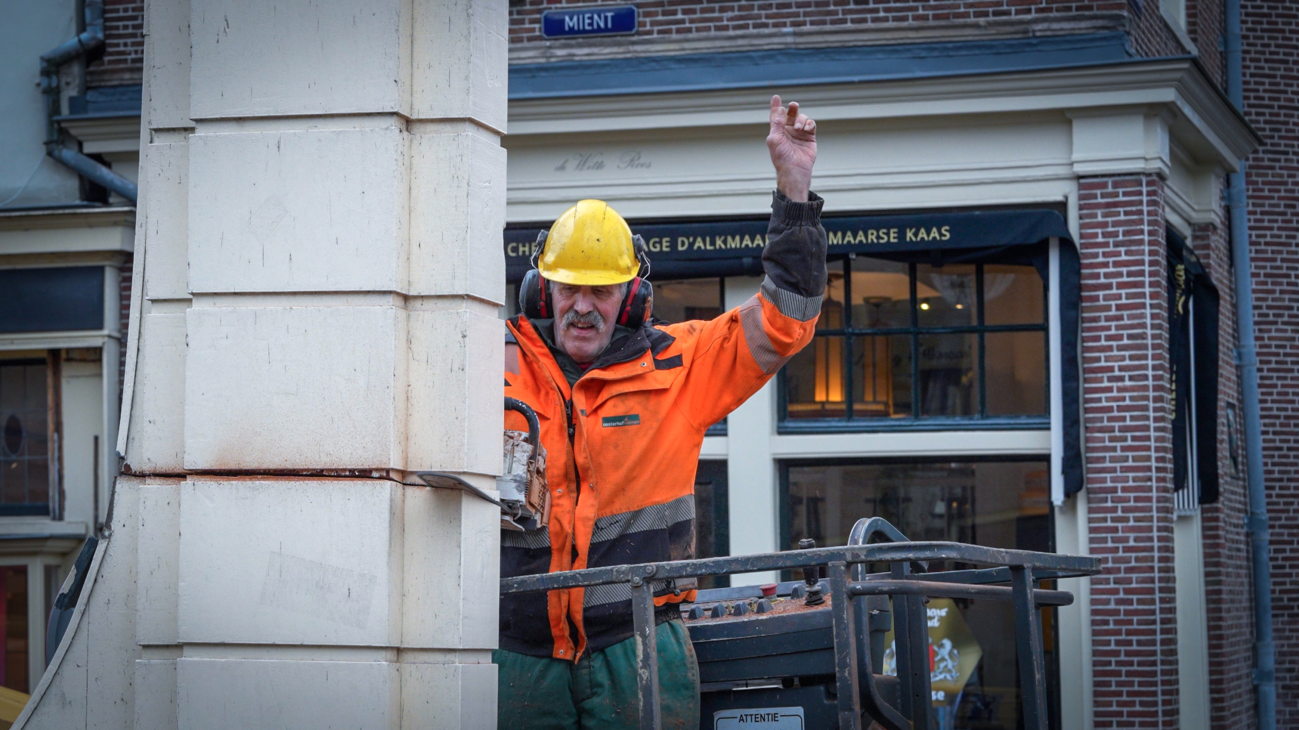 Bouwvakker in oranje jas en gele helm staat op een hoogwerker voor een gebouw.