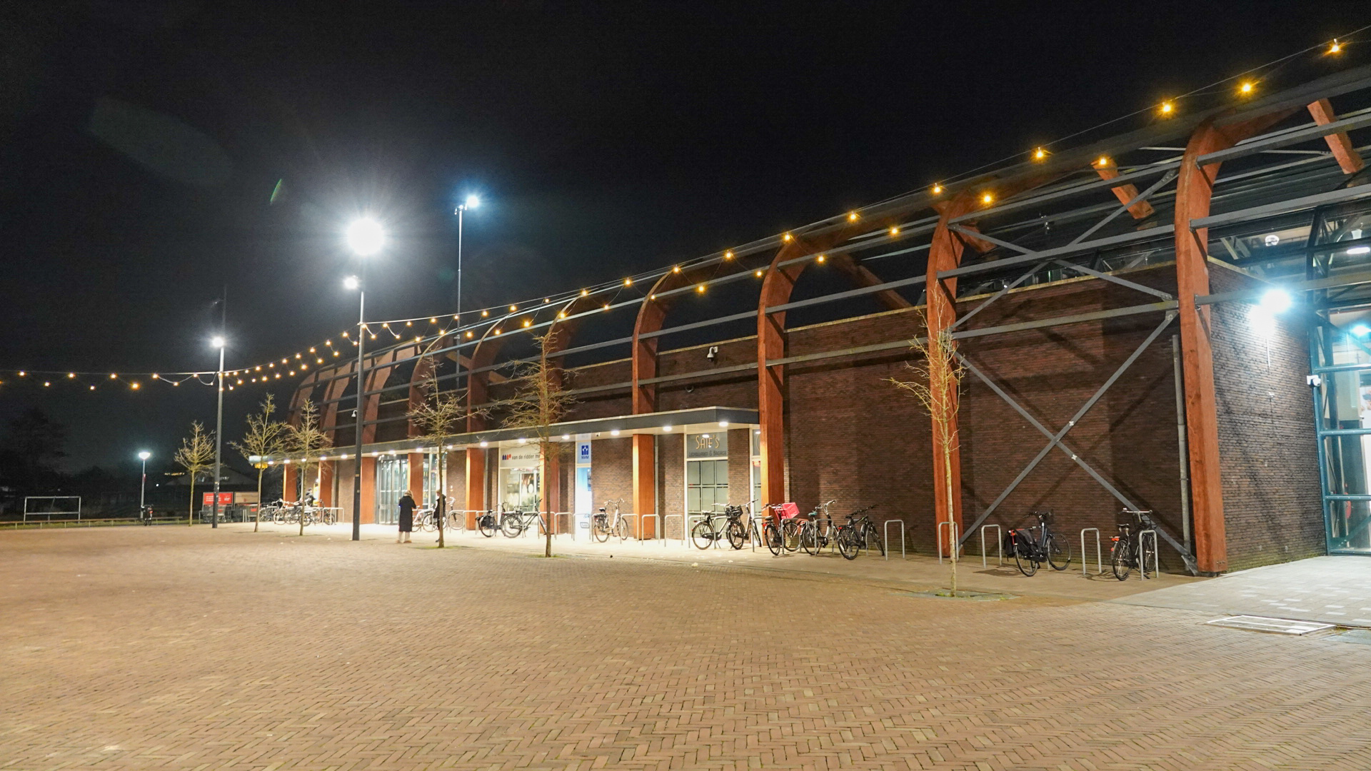 Modern gebouw 's nachts verlicht, met fietsenrekken en enkele fietsen ervoor. Straatverlichting en decoratieve lichtsnoeren zijn te zien.