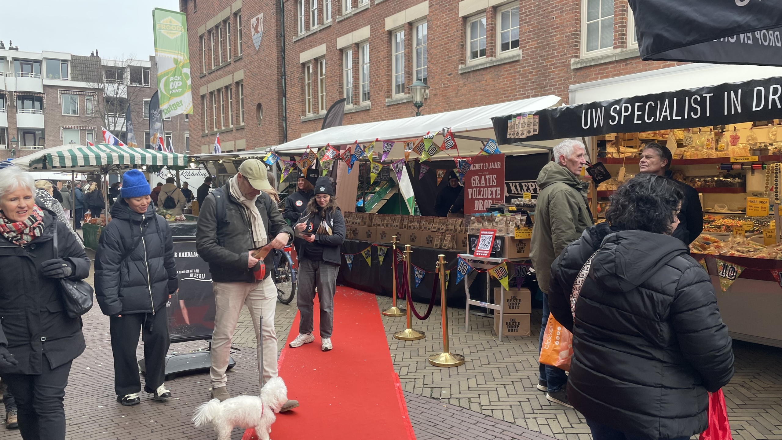 Mensen wandelen langs kraampjes op een markt, enkele personen dragen winterkleding en een witte hond is zichtbaar op een rode loper.