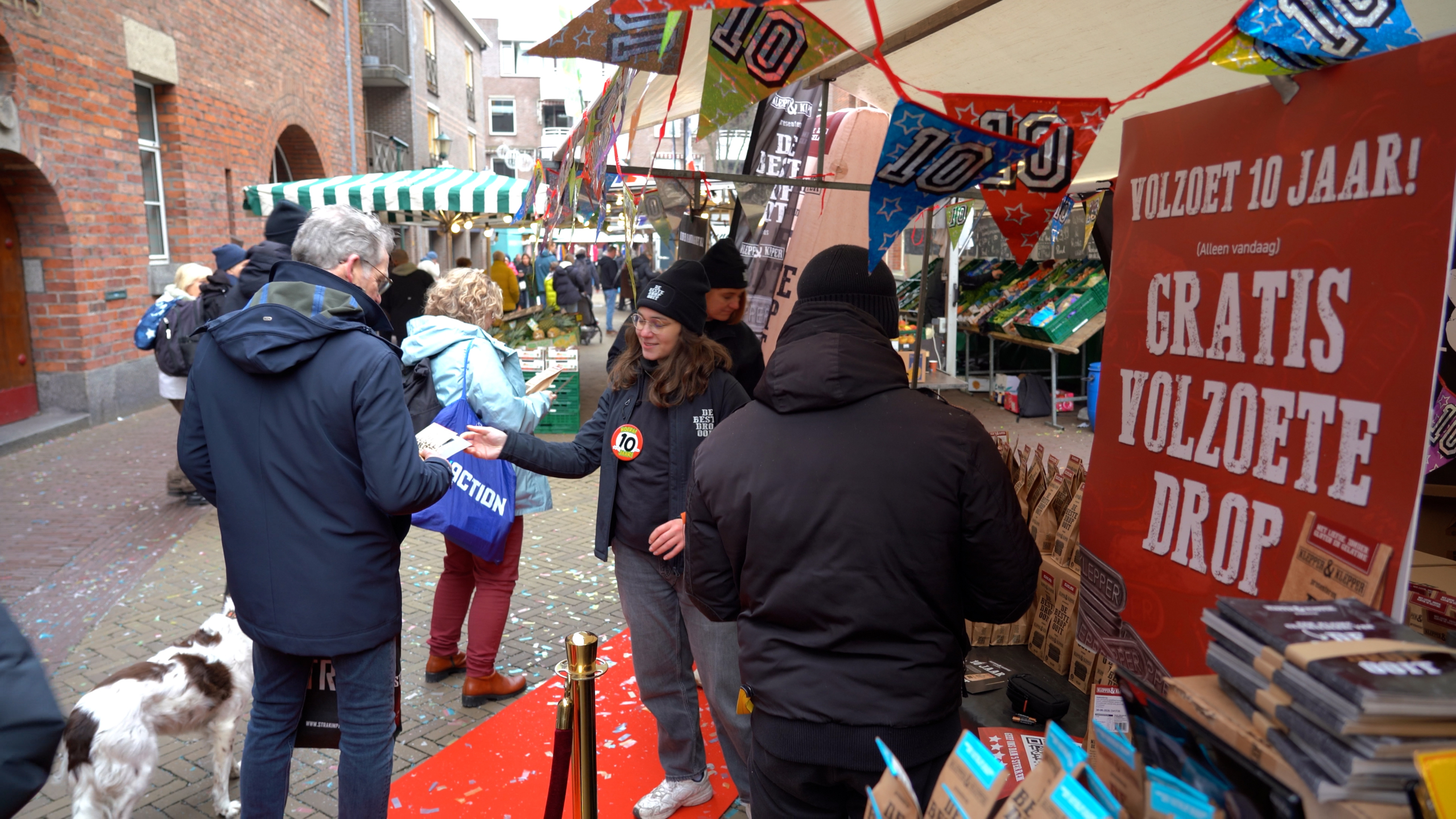 Marktkraam met mensen die gratis volzoete drop uitdelen bij een jubileumviering. Er hangen feestelijke vlaggetjes en er is een bord met de tekst "Volzoet 10 jaar! Gratis volzoete drop".