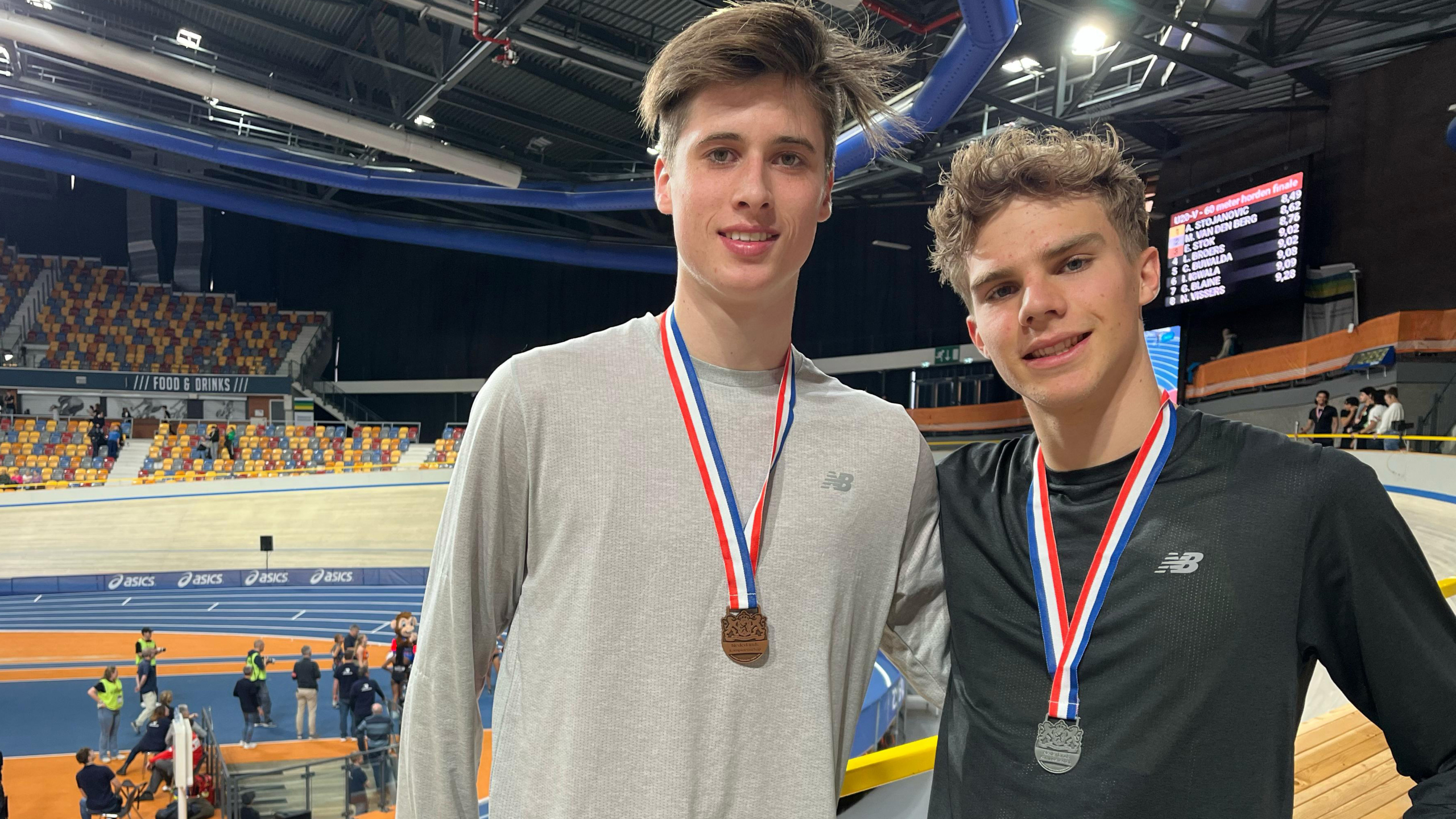 Twee jonge mannen met medailles poseren op een indoor atletiekbaan.