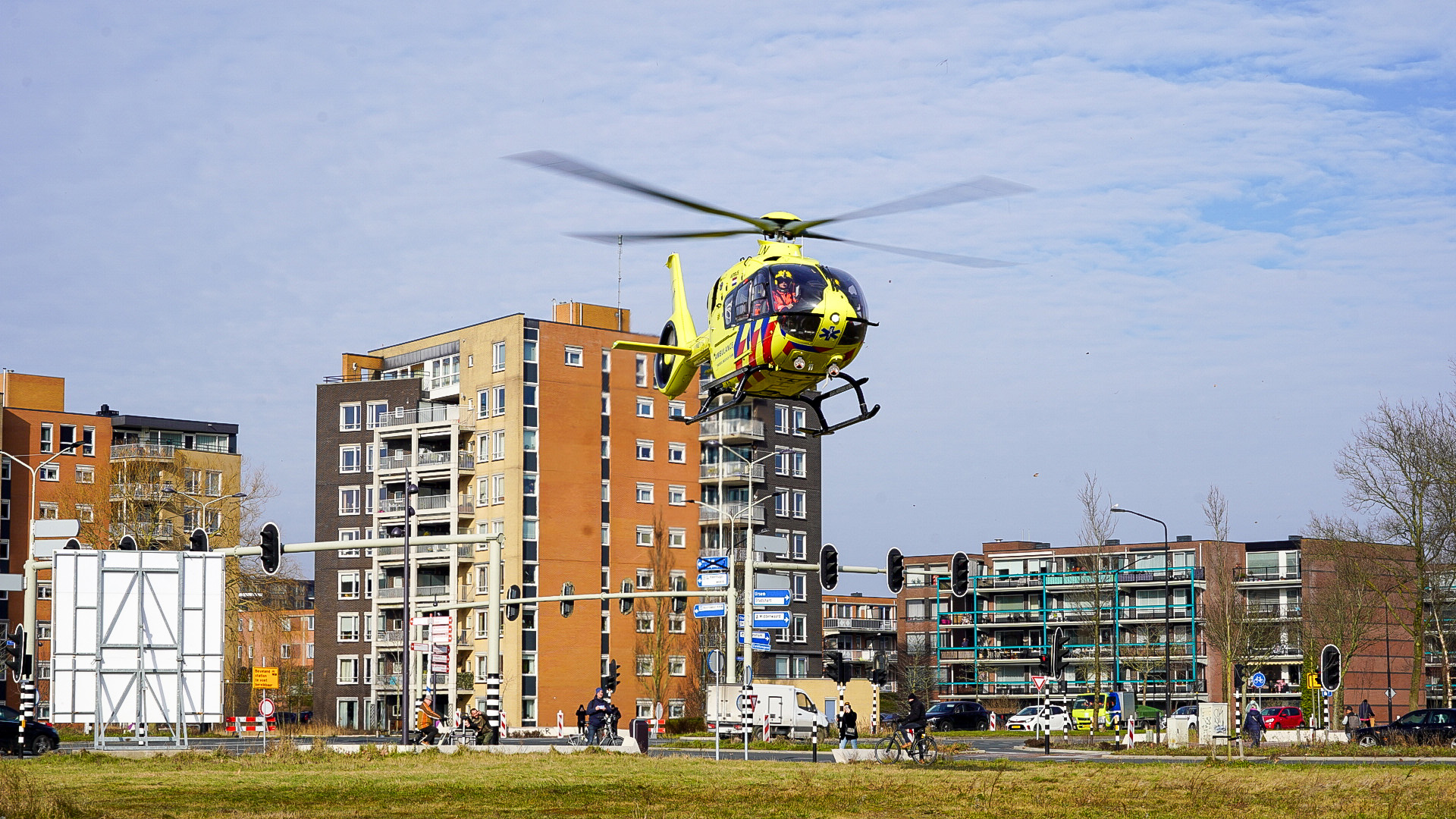 Gele traumahelikopter vliegt laag boven een stedelijk gebied met appartementen en verkeerslichten.