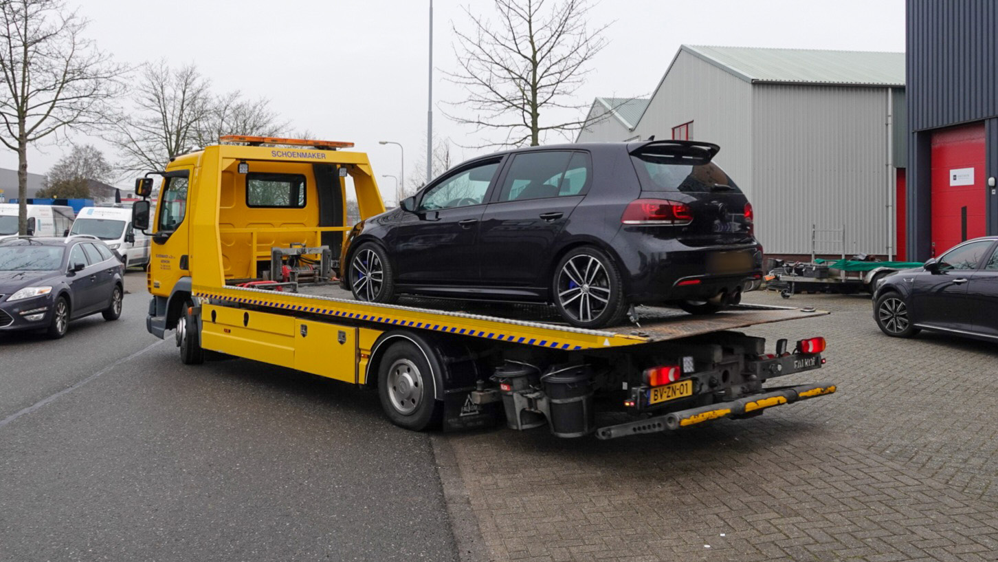 Een zwarte auto op een gele bergingswagen op een straat met geparkeerde en rijdende auto's.