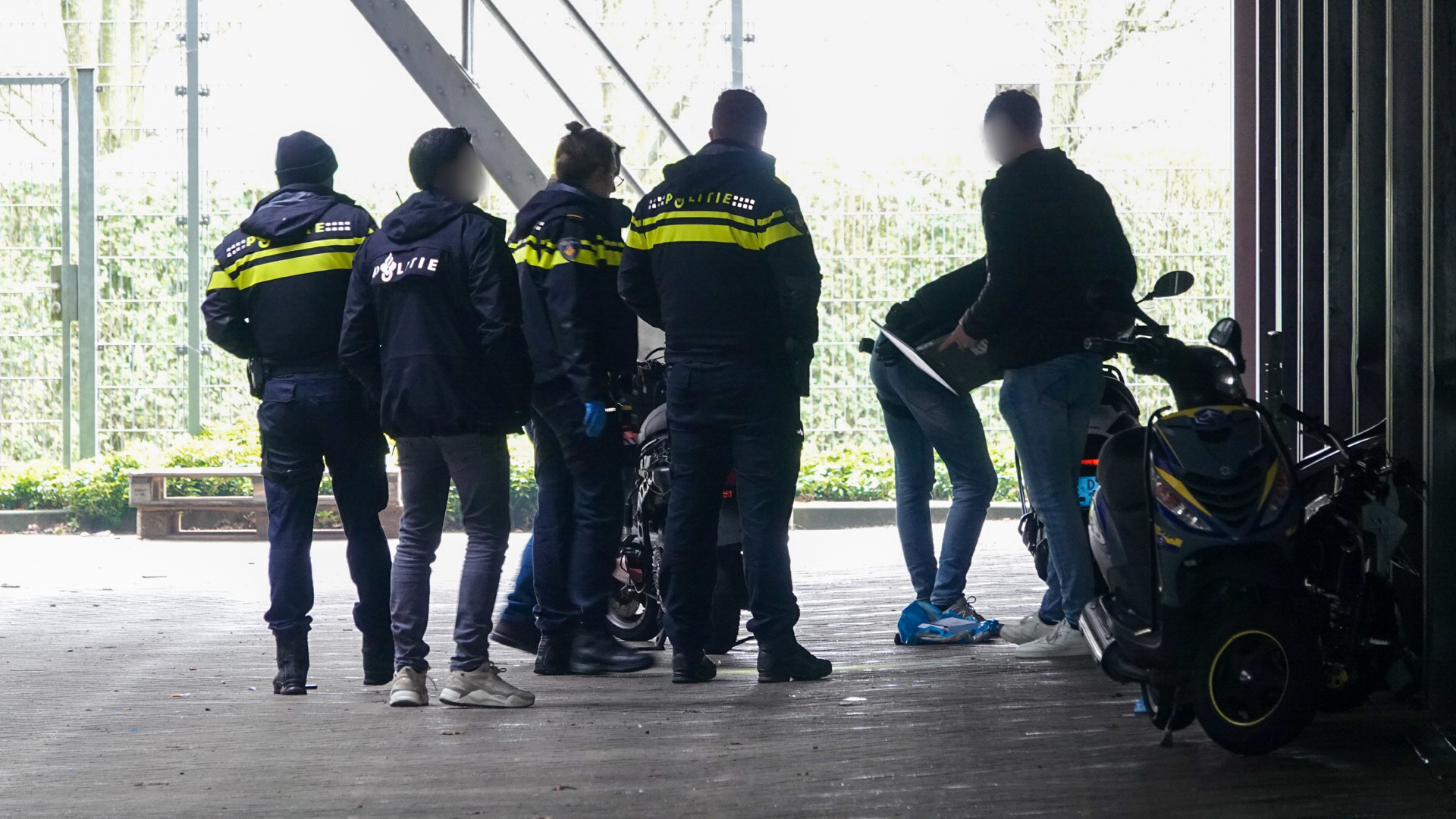 Groep politieagenten omringt een persoon naast een scooter onder een afdak.