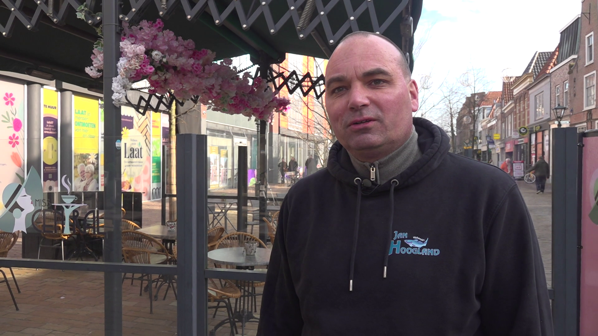 Man in zwarte hoodie staat buiten bij een terras met overkapping en bloemdecoraties. Op de achtergrond winkels in een winkelstraat.