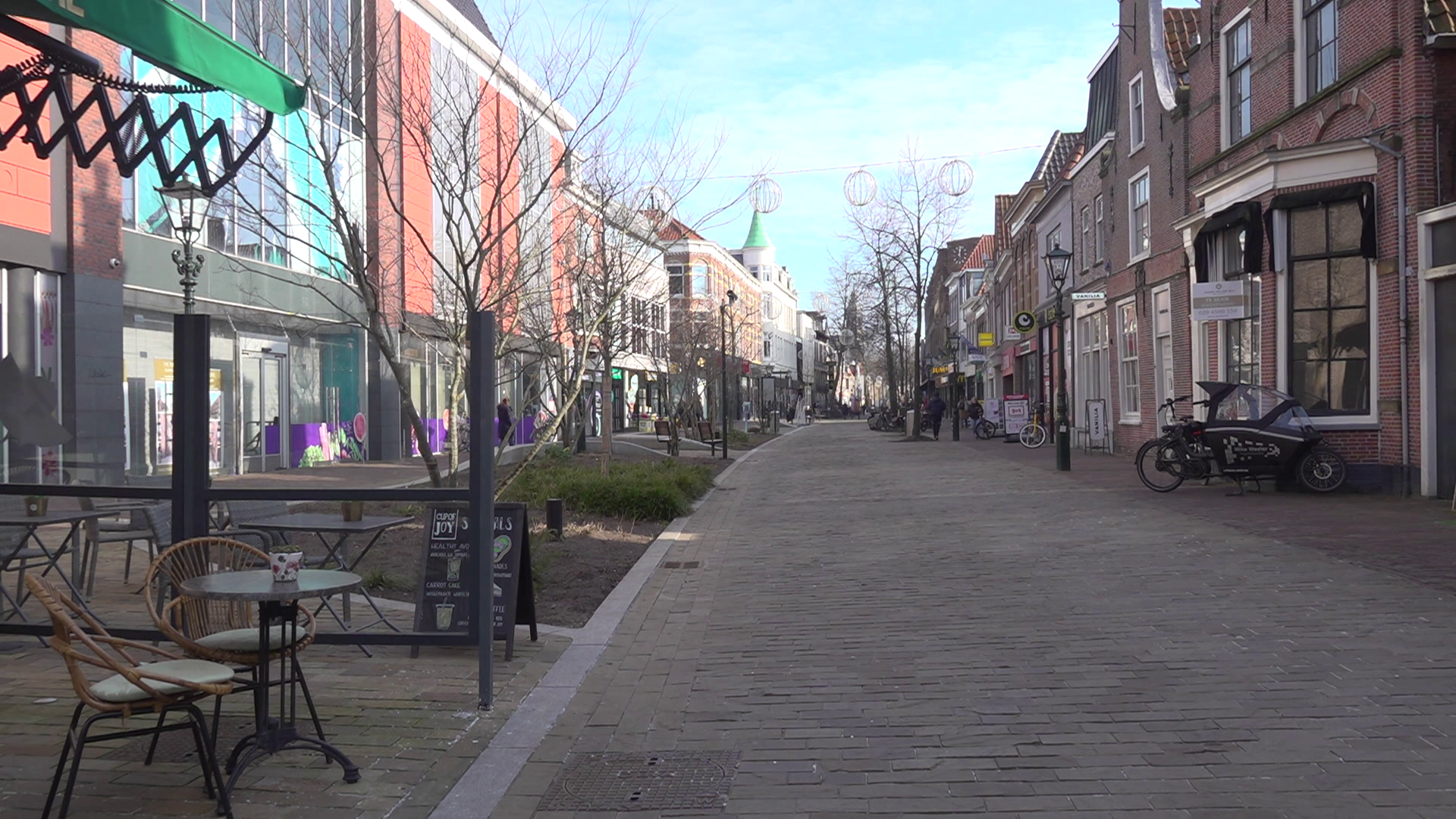 Een lege winkelstraat met terrastafels, fietsen, en huizen aan beide zijden.