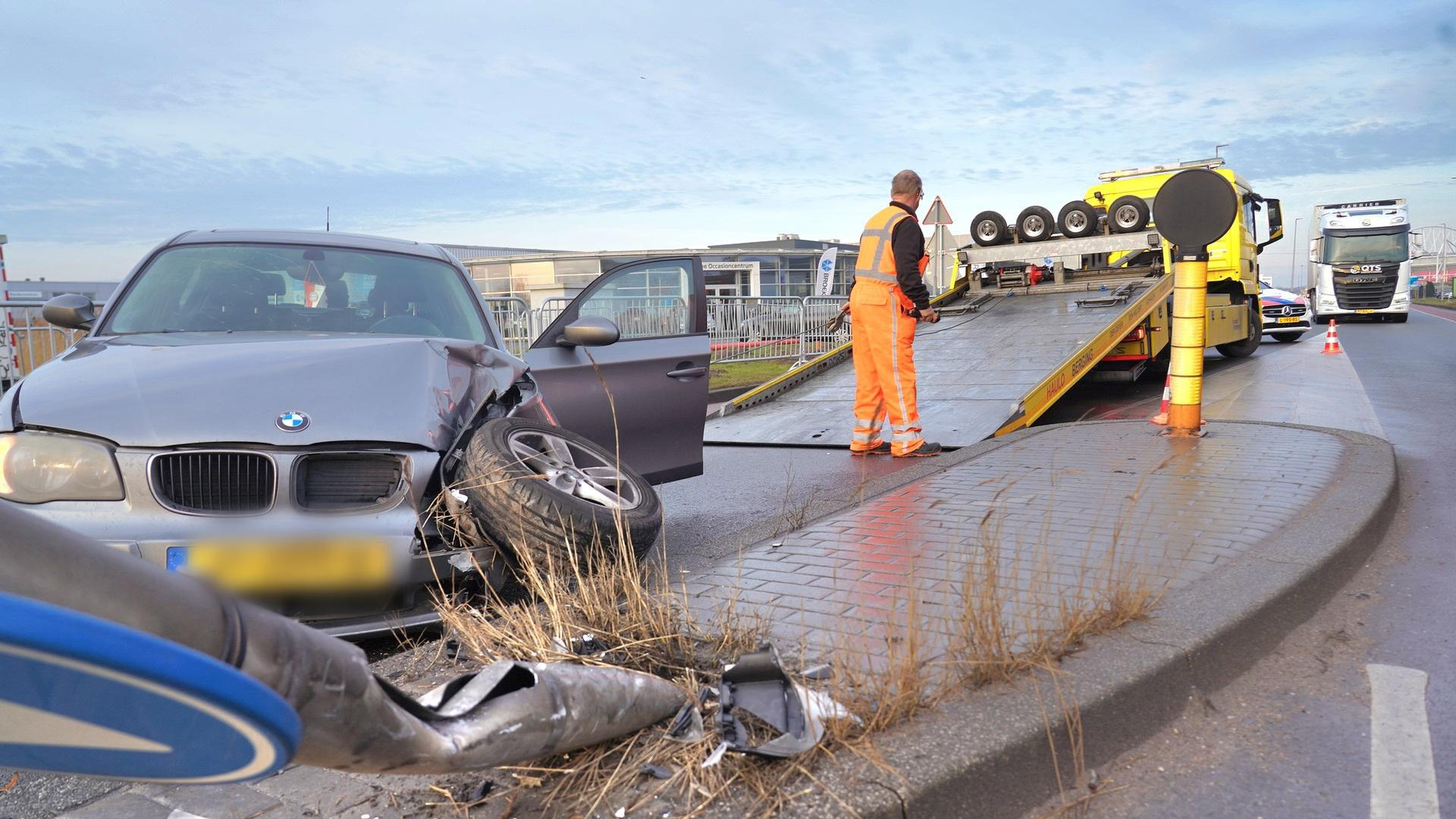 Auto-ongeluk met een beschadigde BMW en een bergingsvoertuig op de achtergrond. Een man in oranje werkkleding staat bij de bergingsvrachtwagen.