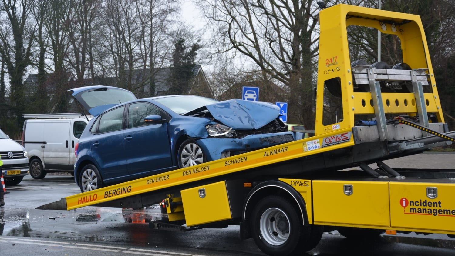 Blauwe auto met schade aan de voorkant op een gele bergingsvrachtwagen, met open motorkap, geparkeerd op een nat wegdek.