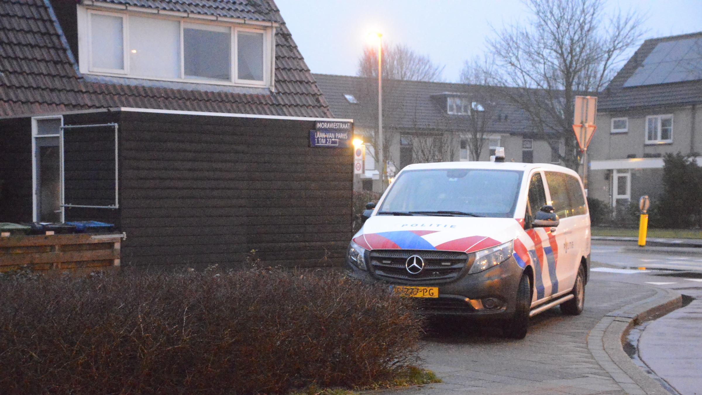 Een politieauto staat geparkeerd in een woonwijk bij een huis met donkere gevelbekleding, naast een straatnaambordje.