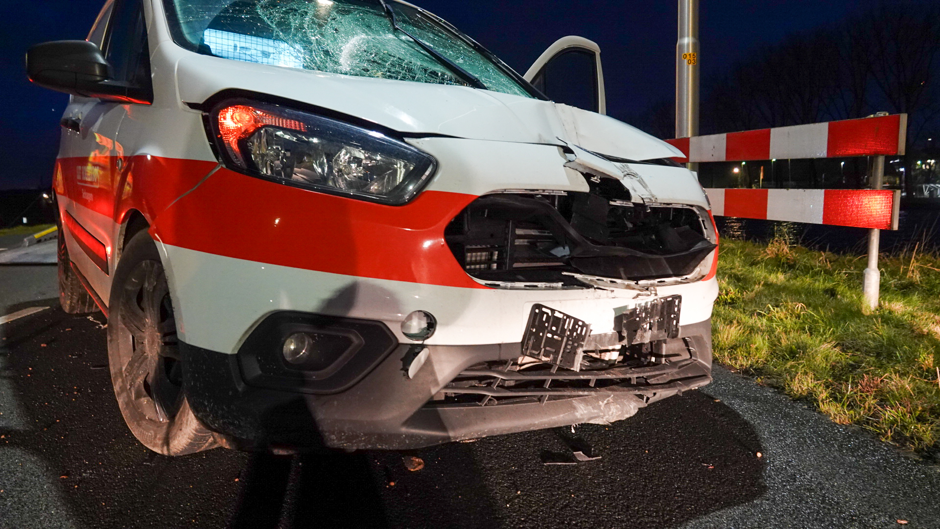 Beschadigde voorkant van een wit met rood voertuig met kapotte bumper en gebarsten voorruit, geparkeerd op een weg bij nacht.