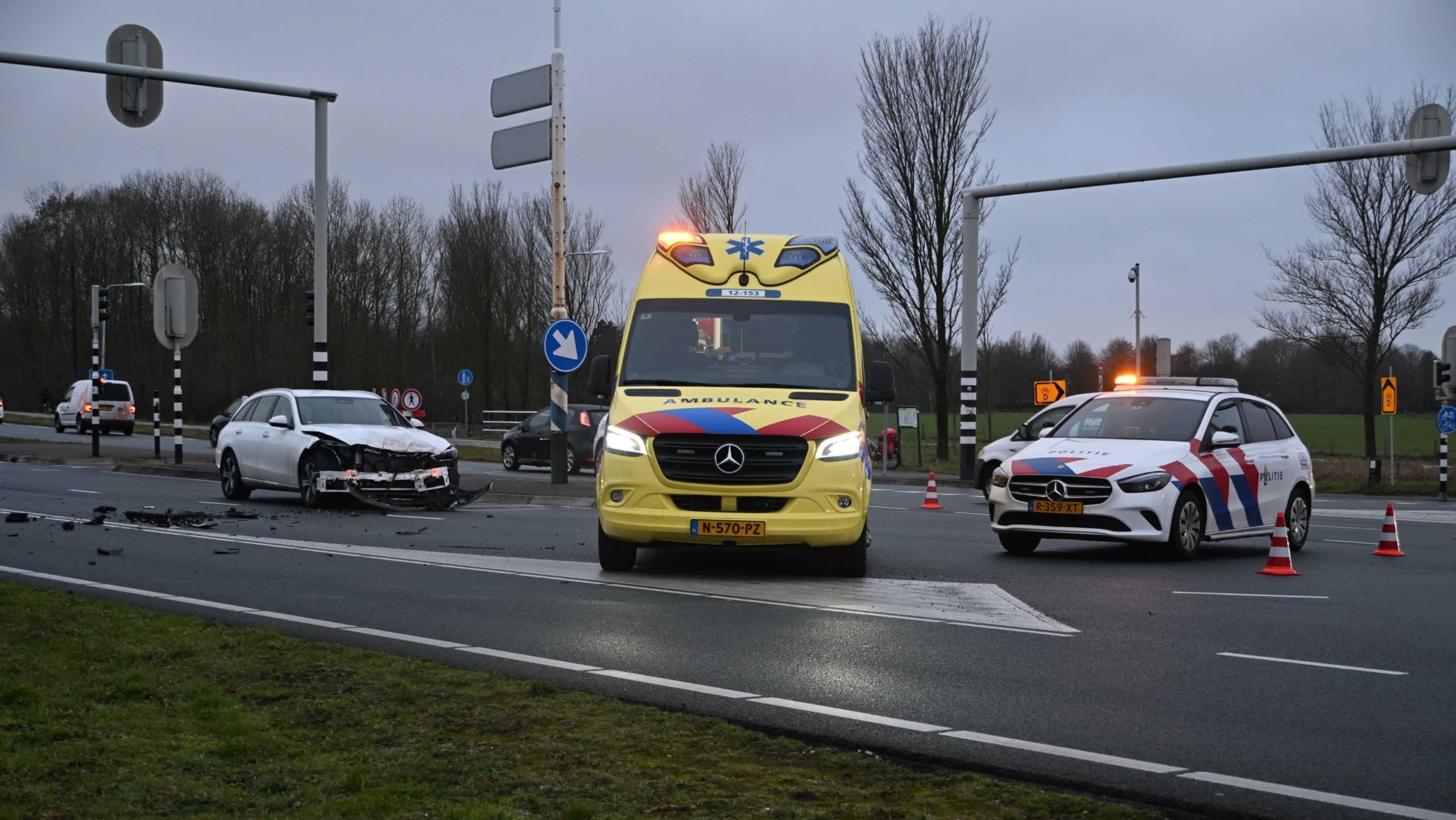 Verkeersongeval met een beschadigde witte auto, een ambulance en een politieauto op een weg met verkeerskegels.