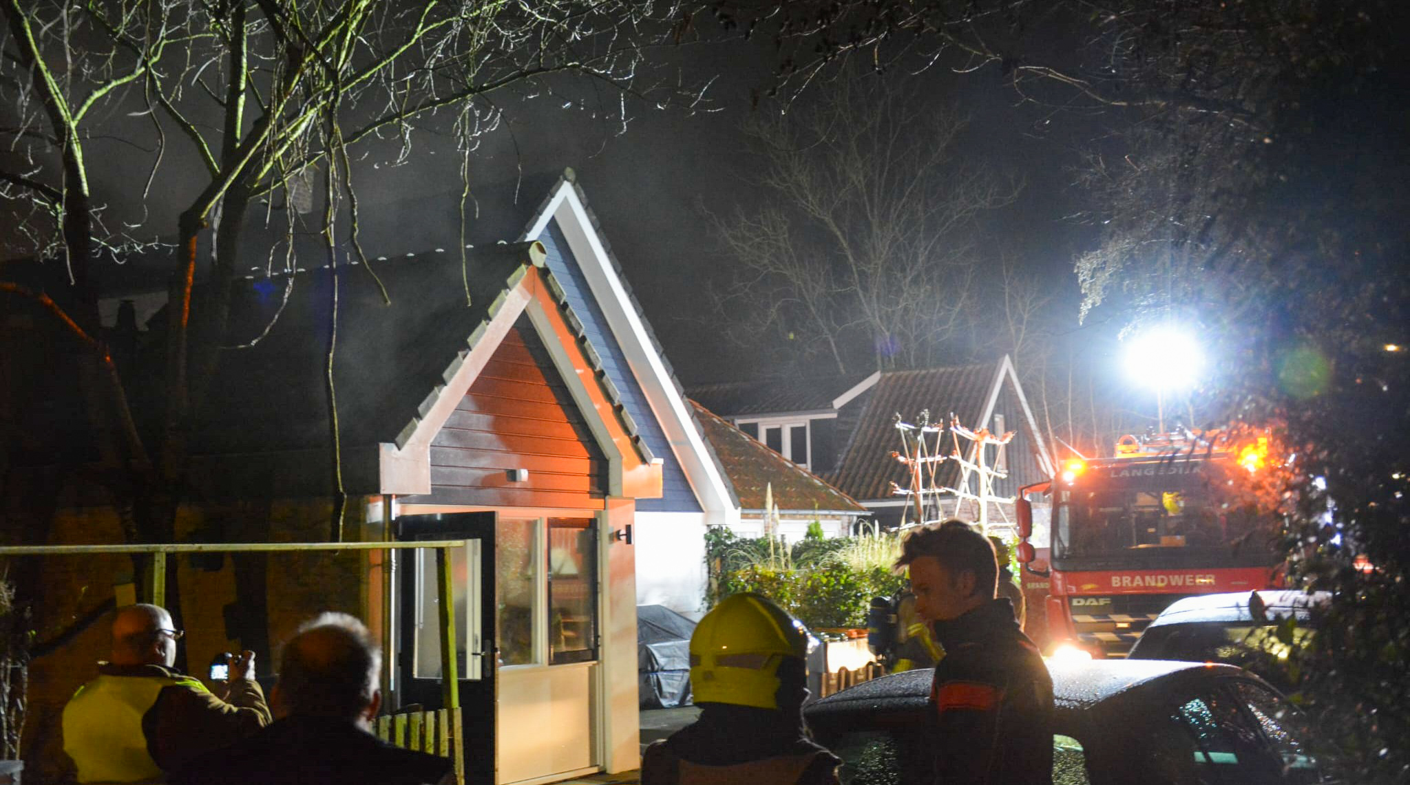 Brandweerauto en brandweerlieden bij nacht bij een huis met lichte rookontwikkeling, omstanders kijken toe.