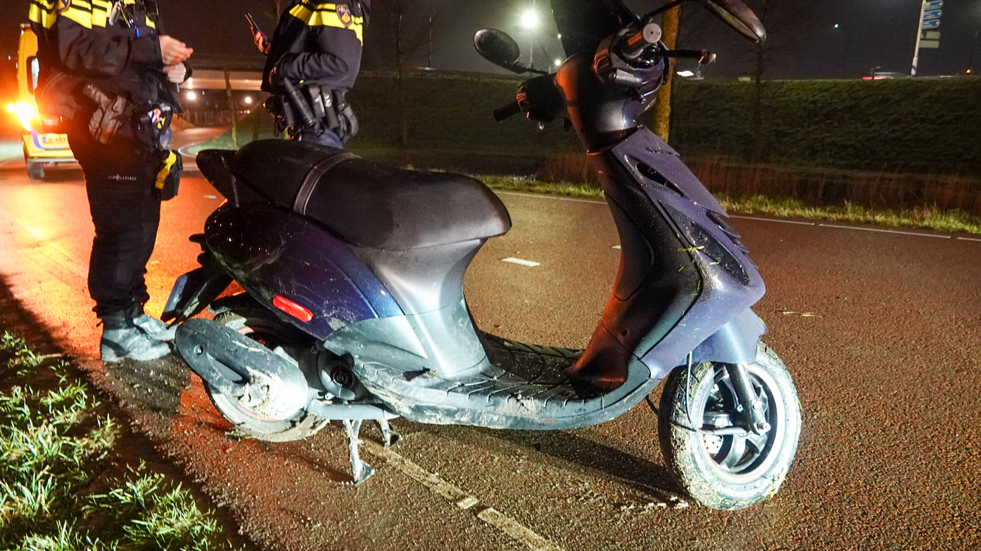 Twee politieagenten staan naast een scooter op een weg 's nachts.