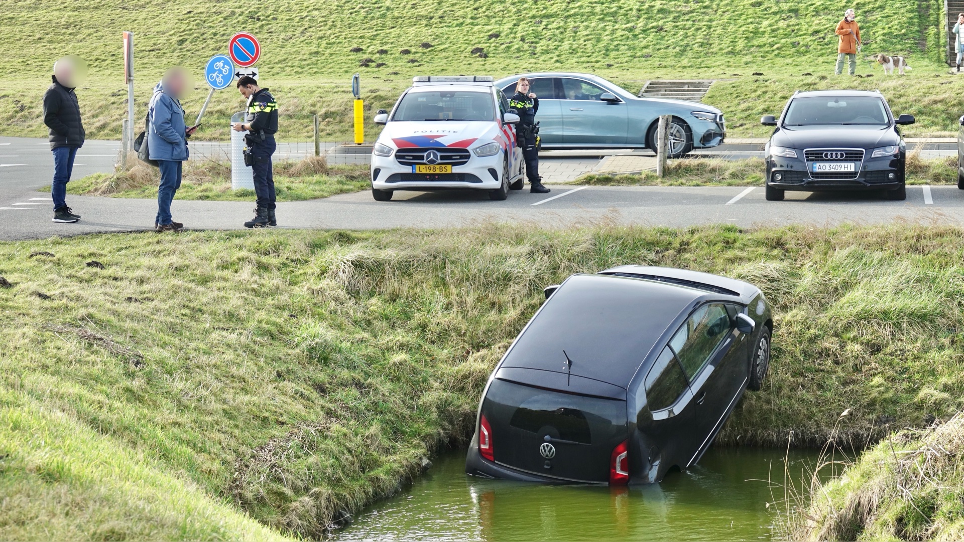 Auto half in een sloot, met politie ter plaatse en mensen die het incident bekijken.