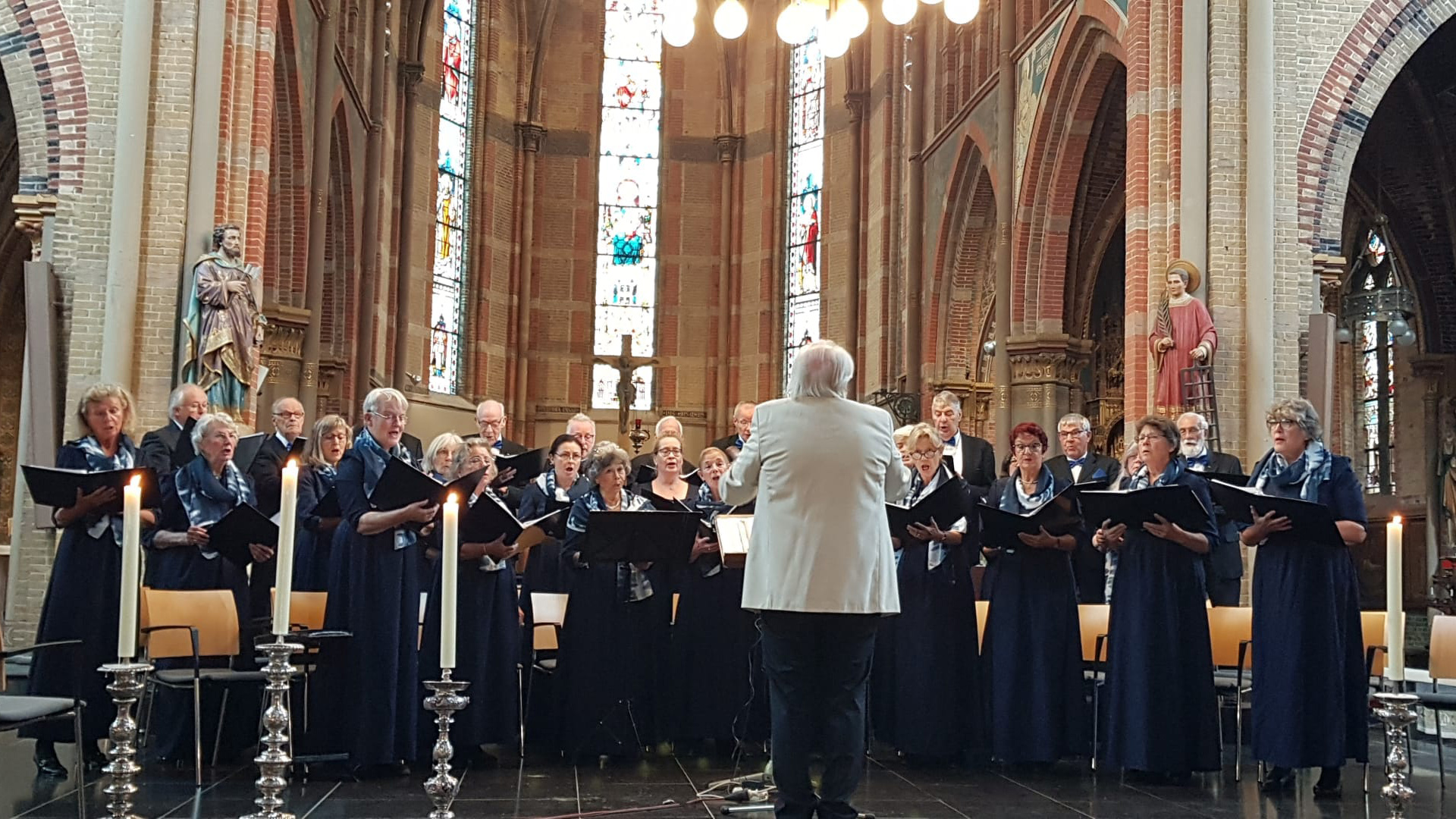 Een koor zingt in een historische kerk met glas-in-loodramen en beelden, geleid door een dirigent in een wit jasje.