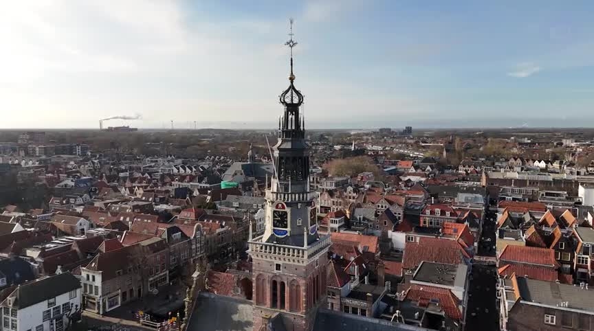 Luchtopname van een oude stad met centraal een historische toren met een klokkentoren en een spitse top, omringd door rode bakstenen huizen en gebouwen.