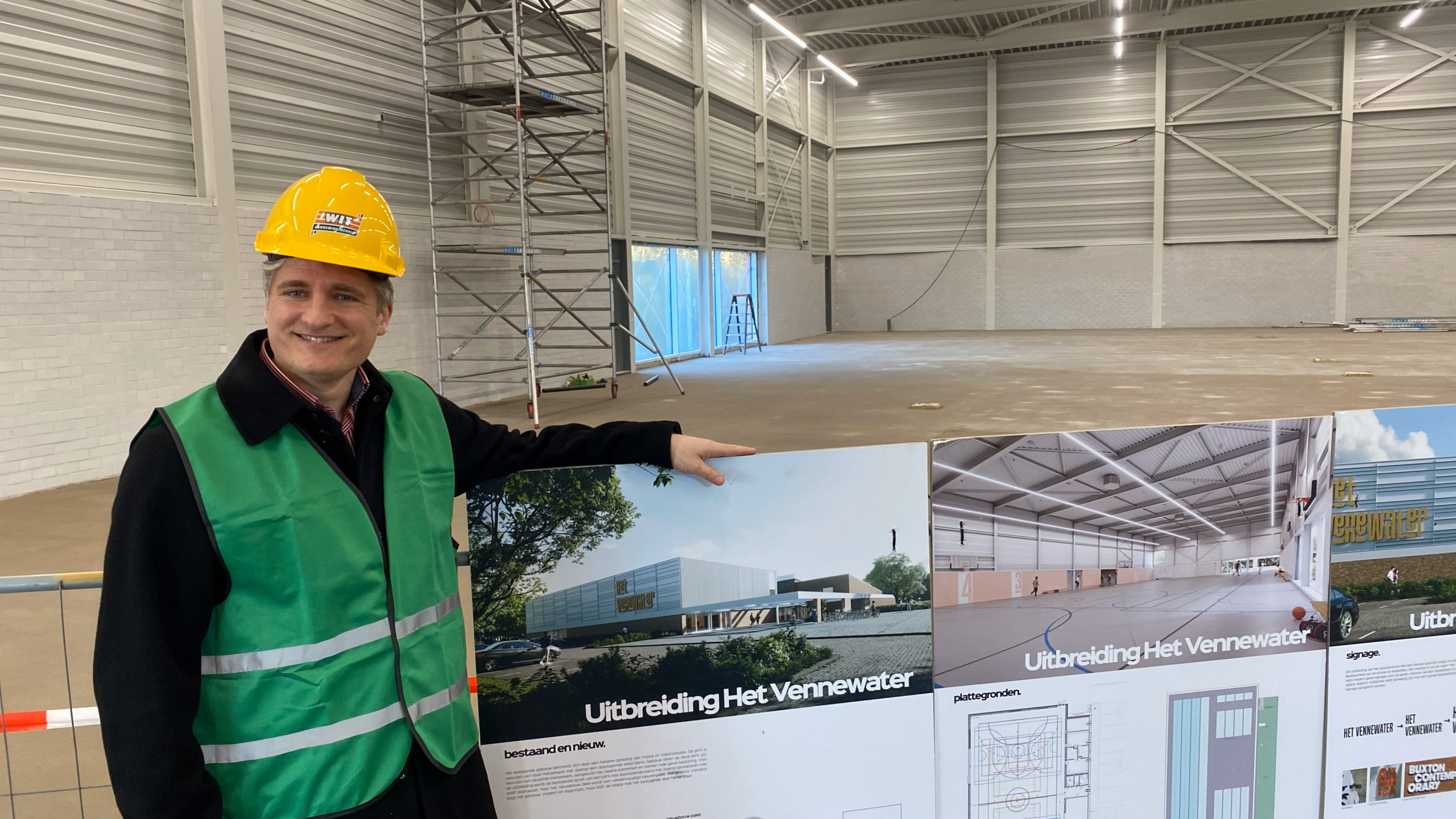 Persoon met gele helm en groene veiligheidshes staat in een bouwplaats, wijzend naar een ontwerp van een gebouw genaamd "Uitbreiding Het Vennewater".